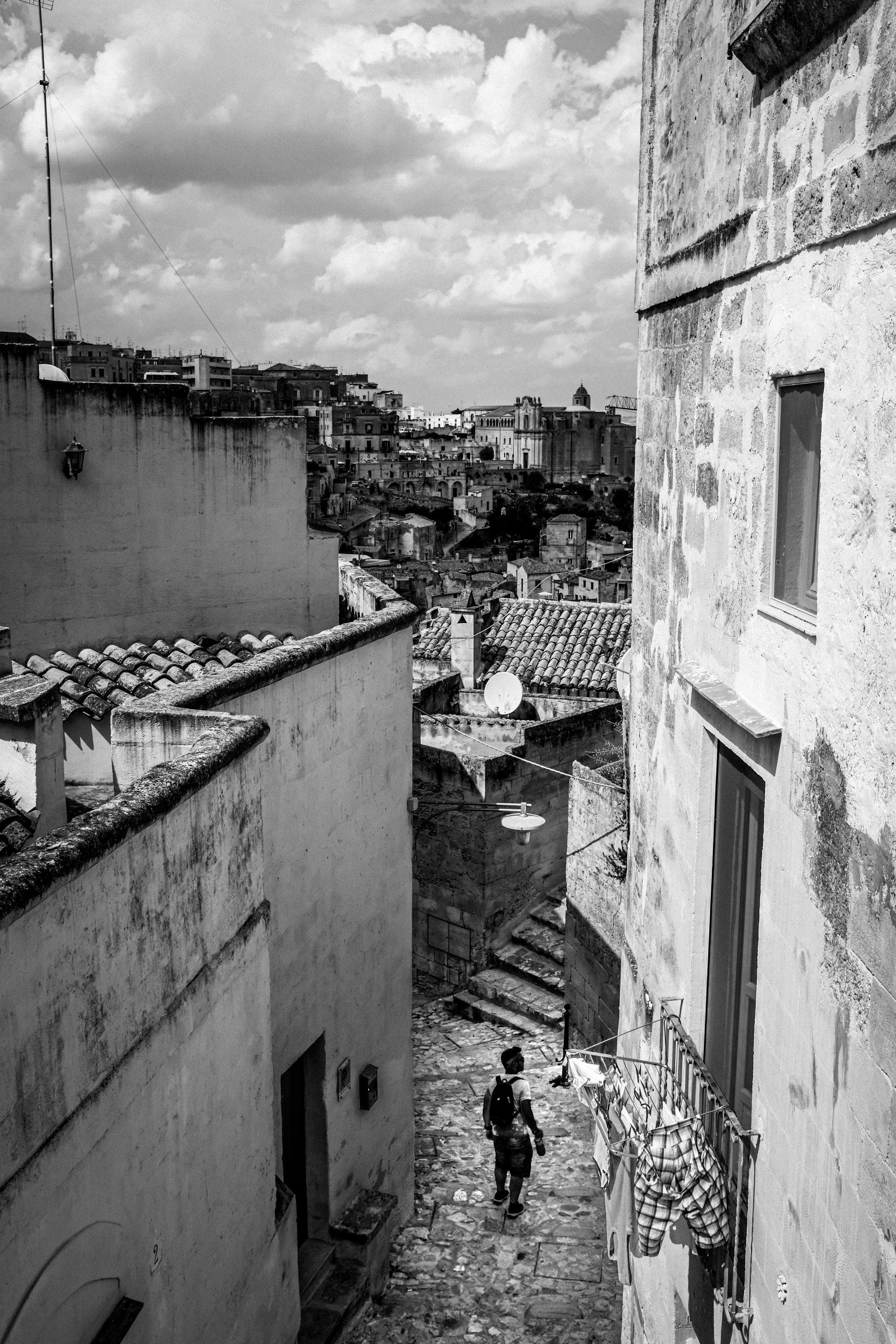 Foto in bianco e nero di un vicolo stretto e in salite con case vecchie e tetti di tegole, una persona vestita casualmente con uno zaino cammina lungo il percorso.
