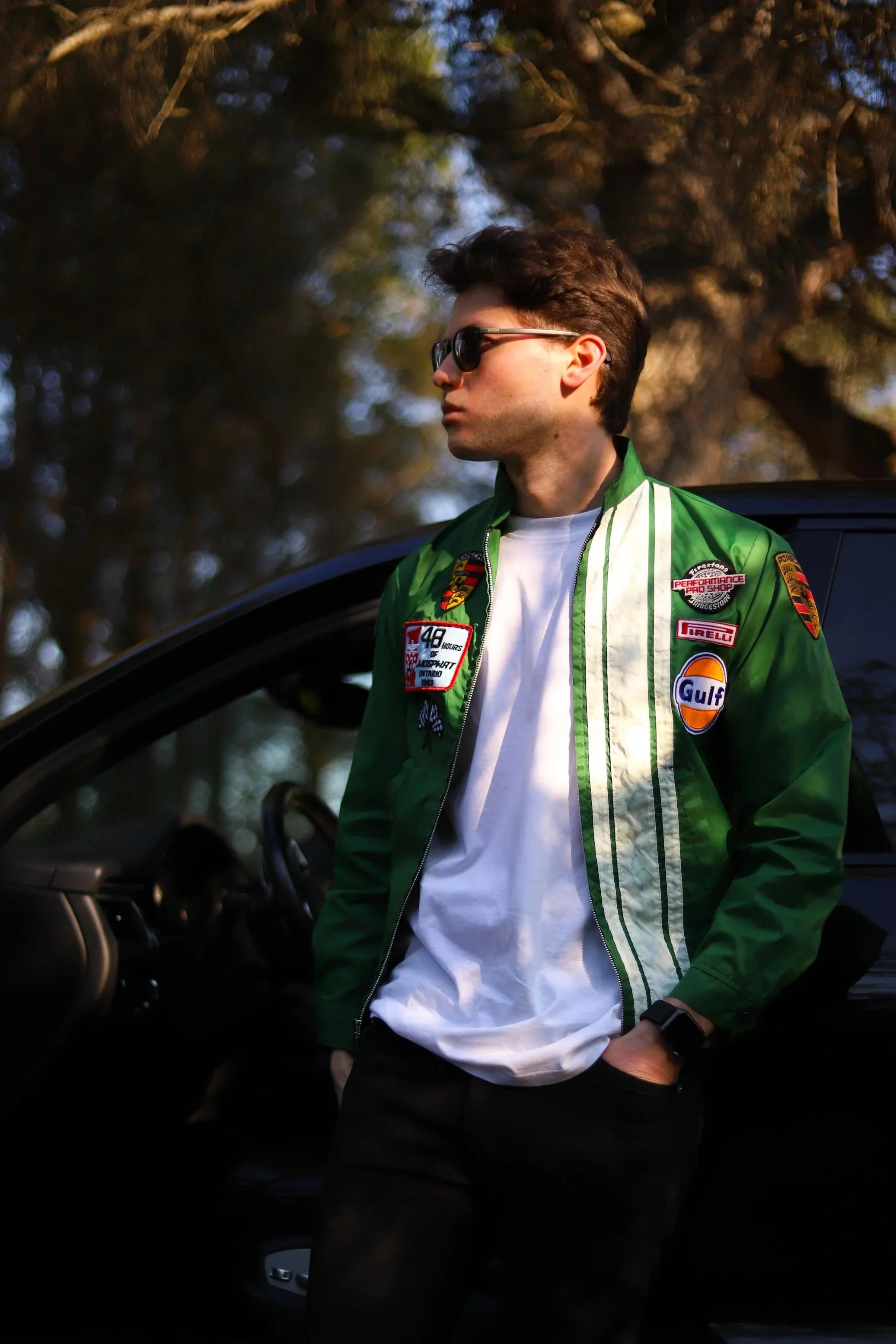 Ragazzo con occhiali da sole che si appoggia a un'auto nera, indossa una giacca verde con patch e una maglietta bianca, in un ambiente all'aperto con alberi in sfondo.