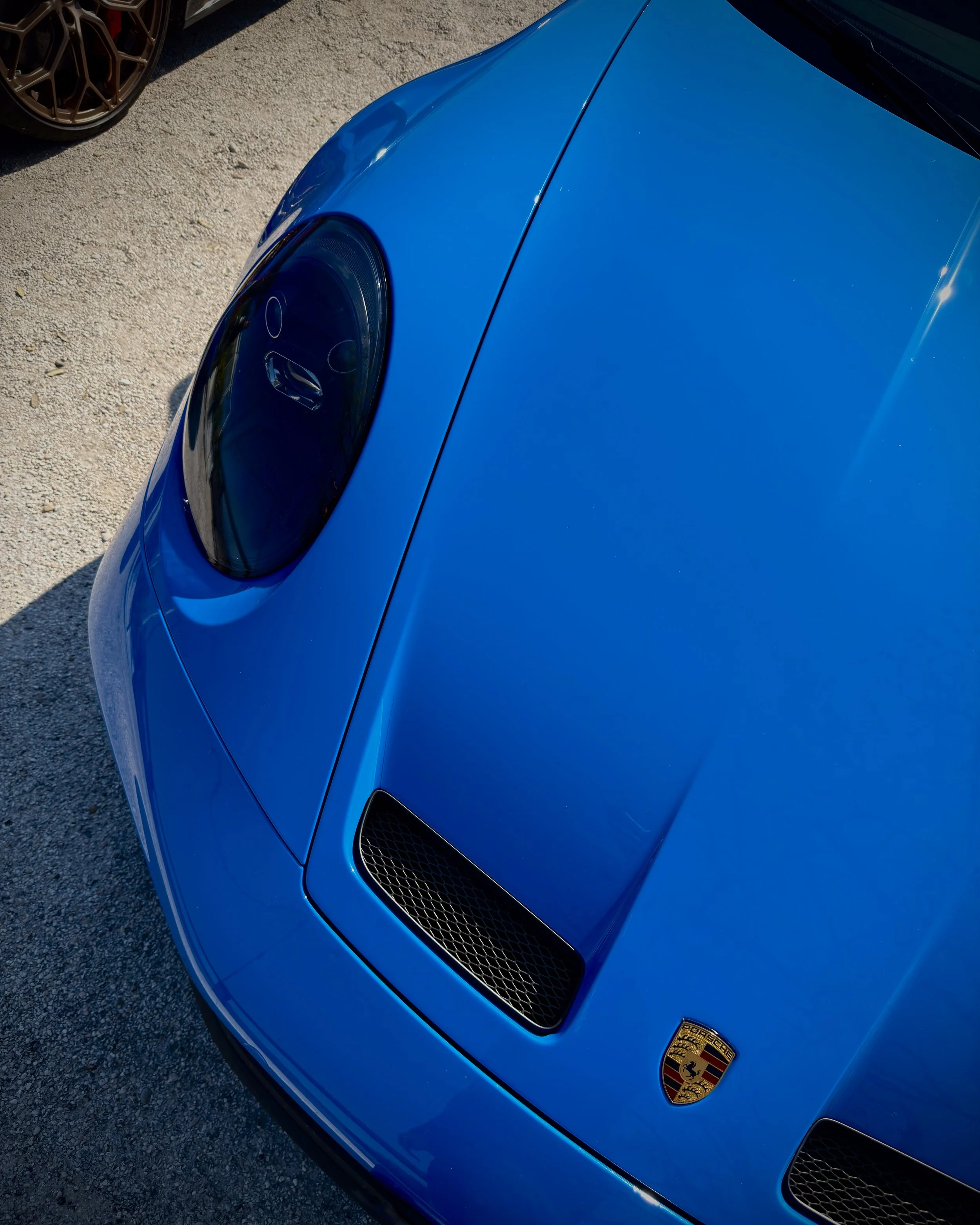 Dettaglio di una vettura sportiva Porsche di colore blu, vista dalla parte anteriore sinistra. Viene mostrato il cofano motore, la griglia dell’aria e il logo Porsche.