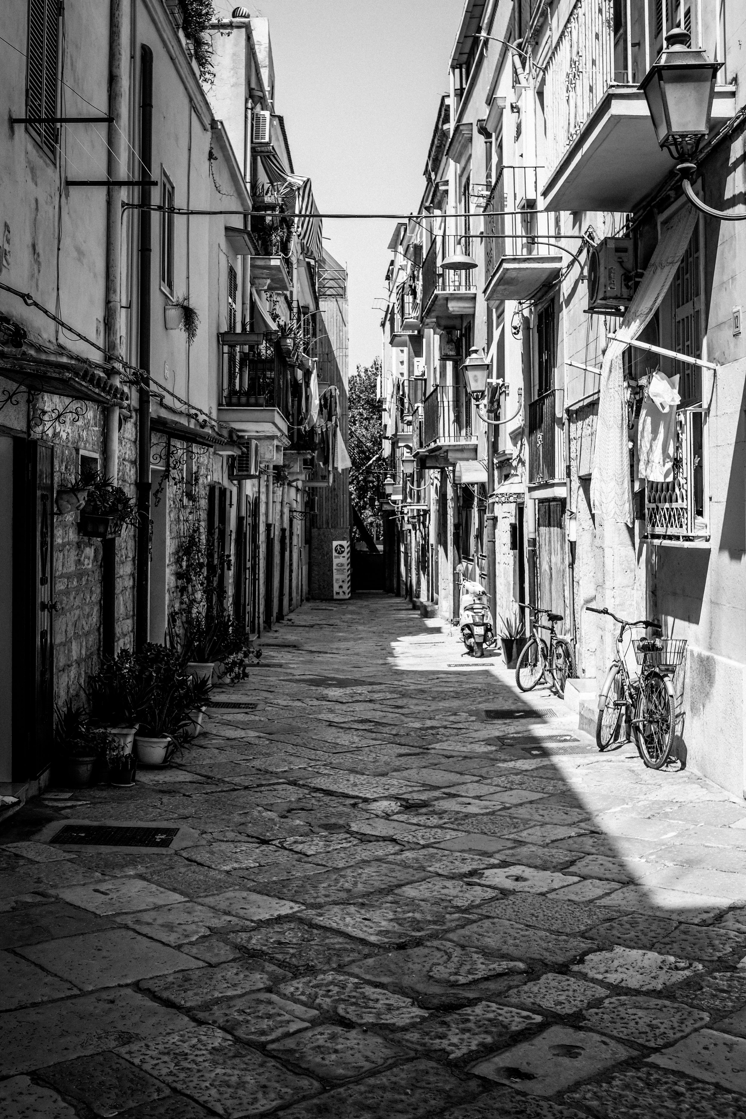 Una strada acciottolata in un quartiere residenziale con case a più piani, alcune con piante in vaso e biciclette parcheggiate. Ombra sulla strada e lampioni appesi alle pareti.
