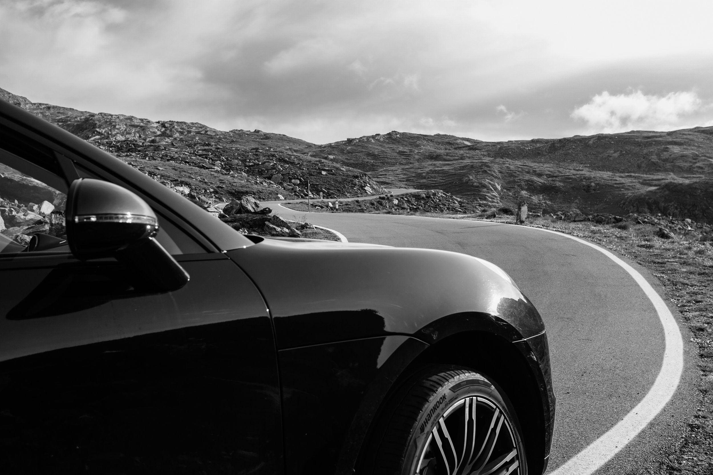 Vista di un'auto nera in curva su una strada di montagna con paesaggio roccioso e cielo nuvoloso.
