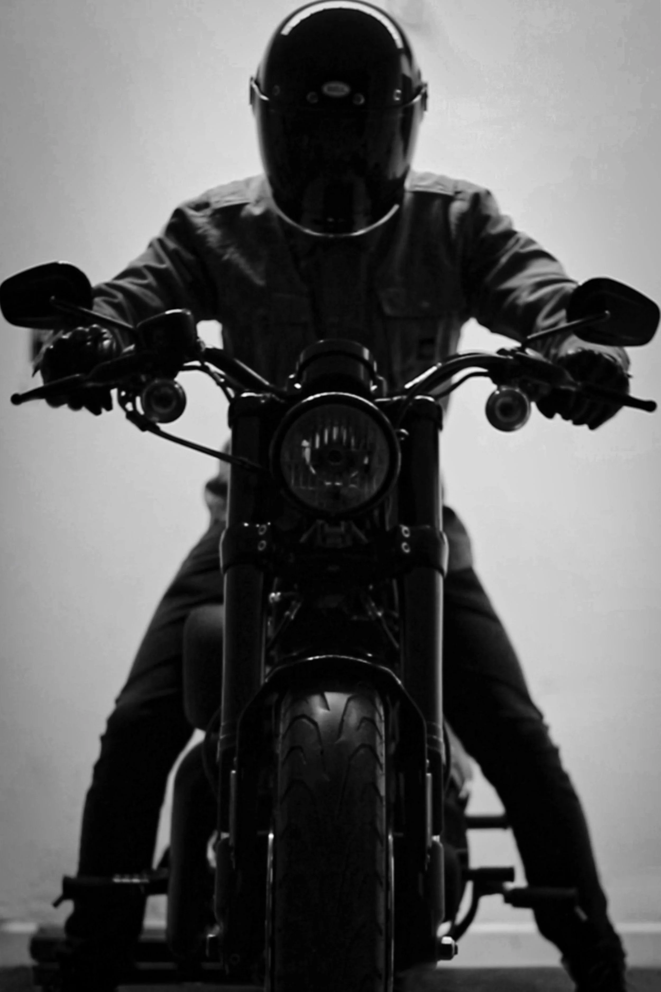 Immagine in bianco e nero di una persona indossando casco e giacca, seduta su una motocicletta, vista frontalmente.