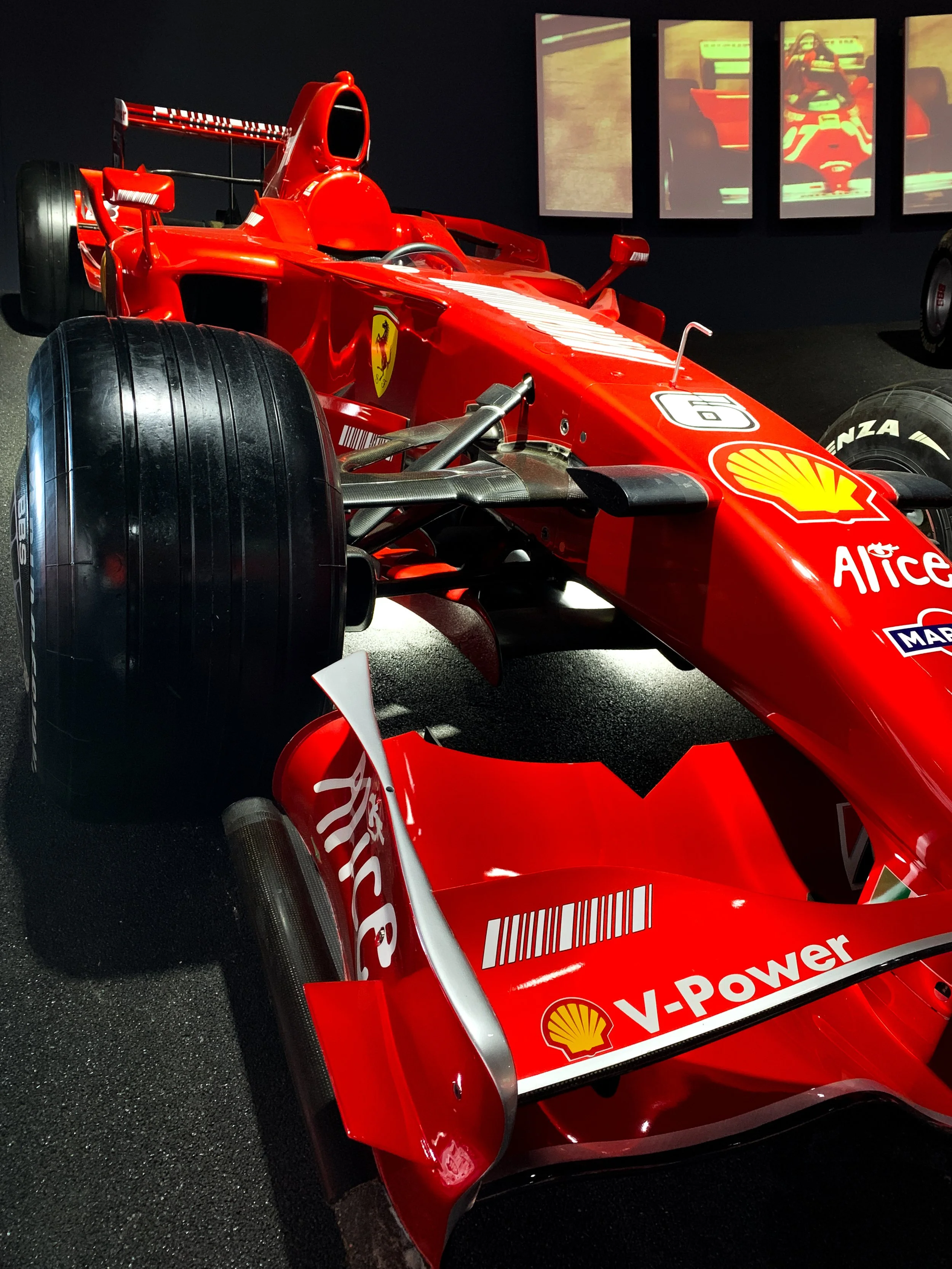 Una vettura da corsa Ferrari di Formula 1 esposta al museo, rossa con loghi sponsor e pneumatici slick. Sullo sfondo, schermi con immagini di auto da corsa.