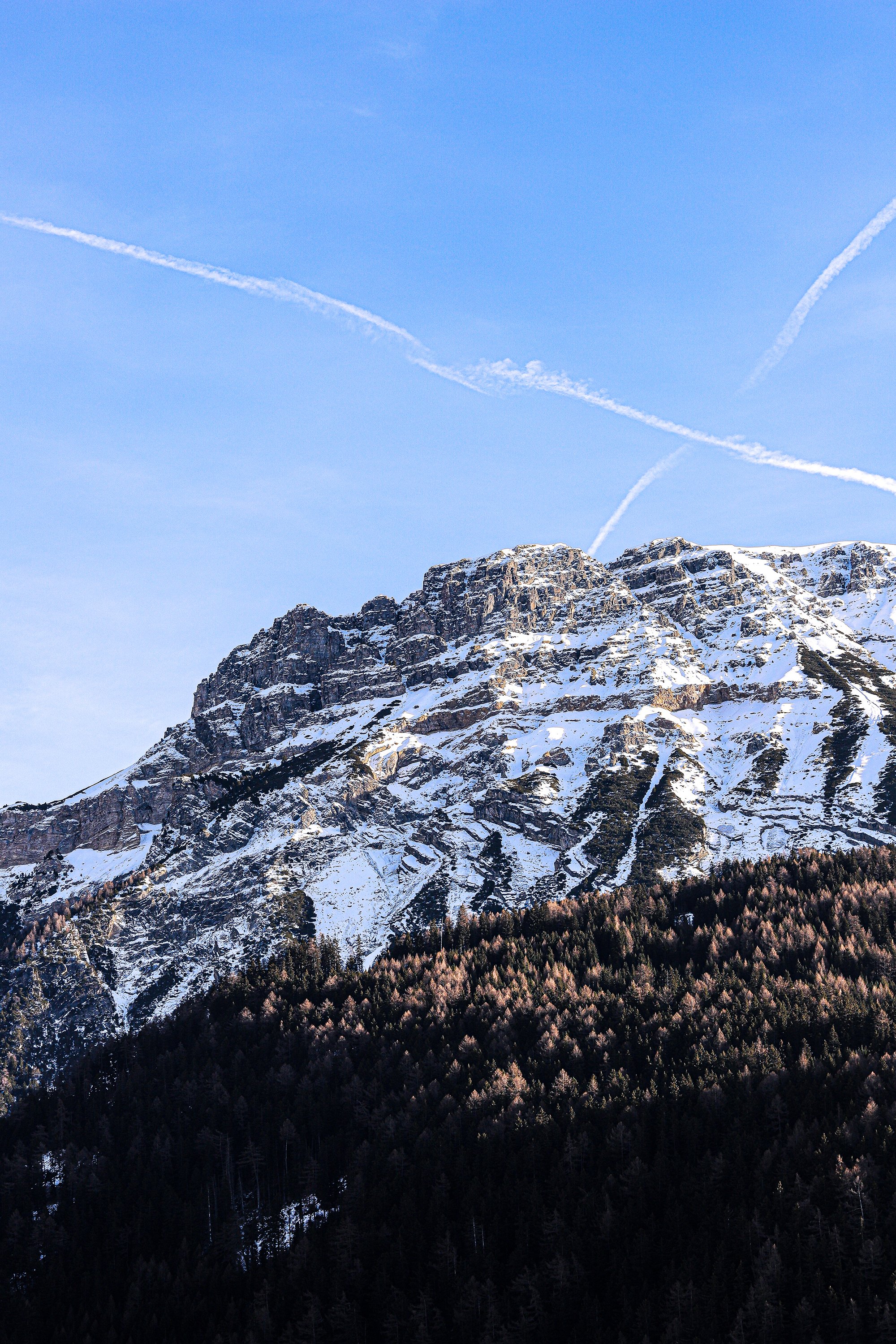 Montagna con cime rocciose e neve, cielo blu con nuvole a forma di linee e alcune tracce di aerei.