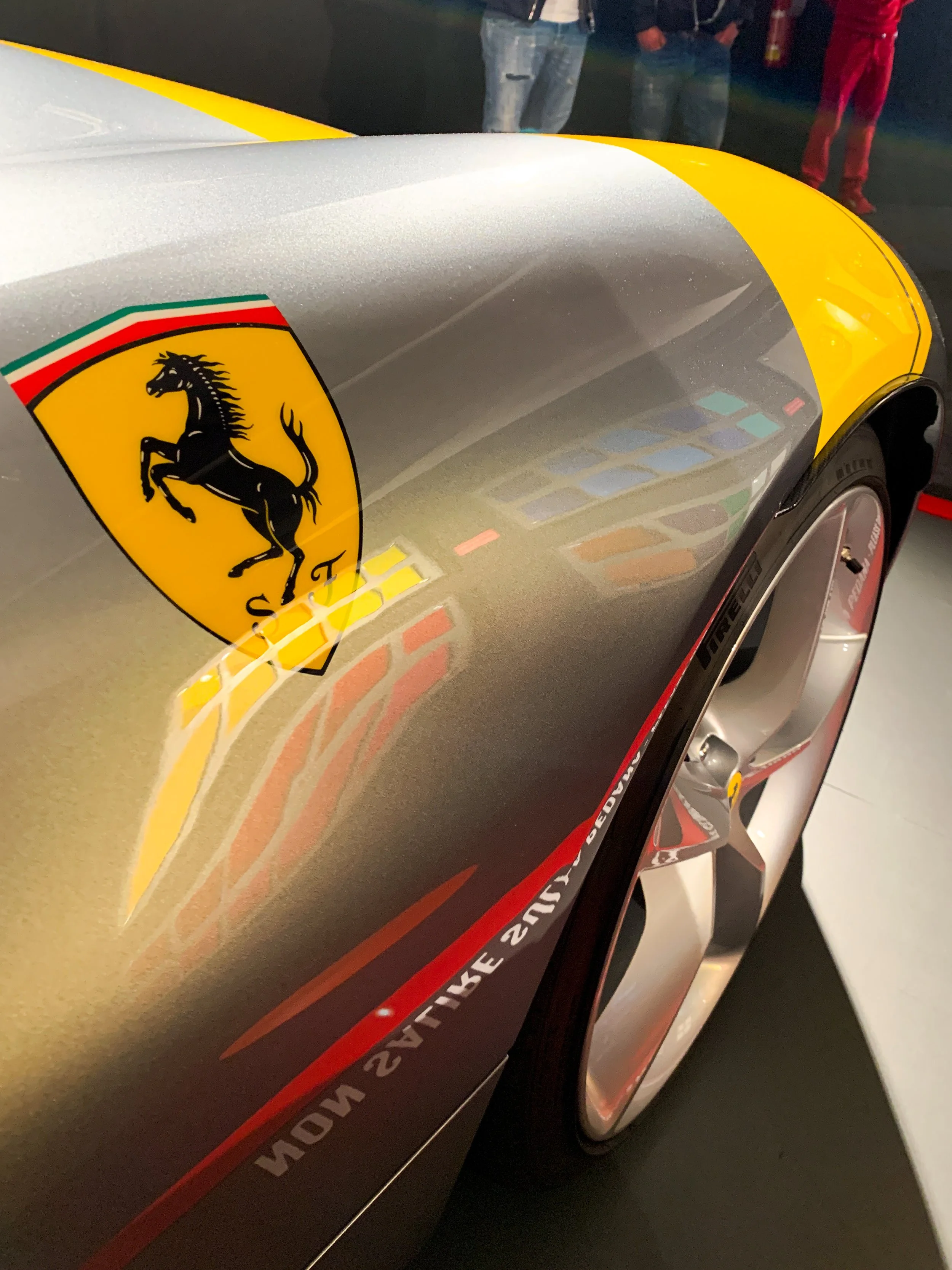 Dettaglio di una vettura di Formula 1 Ferrari con il logo del cavallino rampante, riflessi di luci sui scivoli e i pneumatici, con alcune persone sullo sfondo.