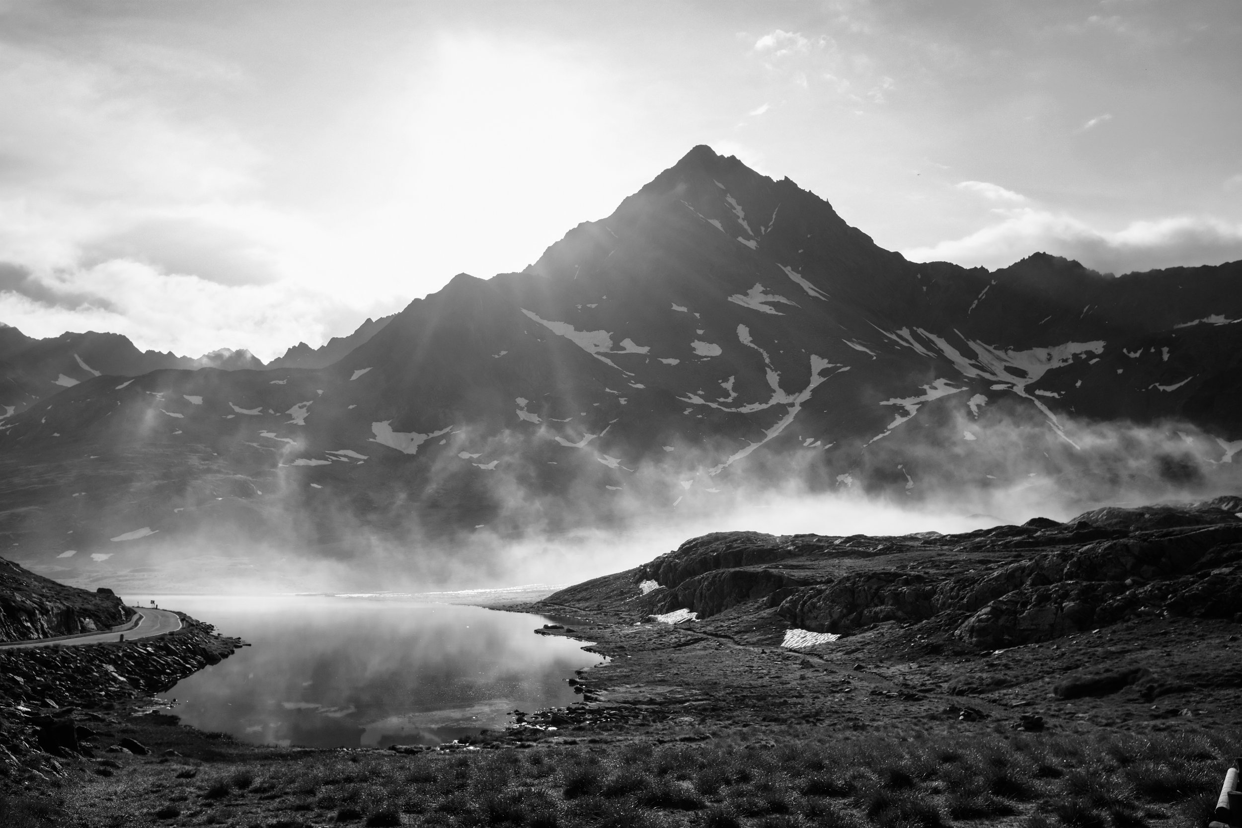 Paesaggio montano con alta cima, lago in primo piano, nebbia e nuvole nel cielo, scena in bianco e nero.