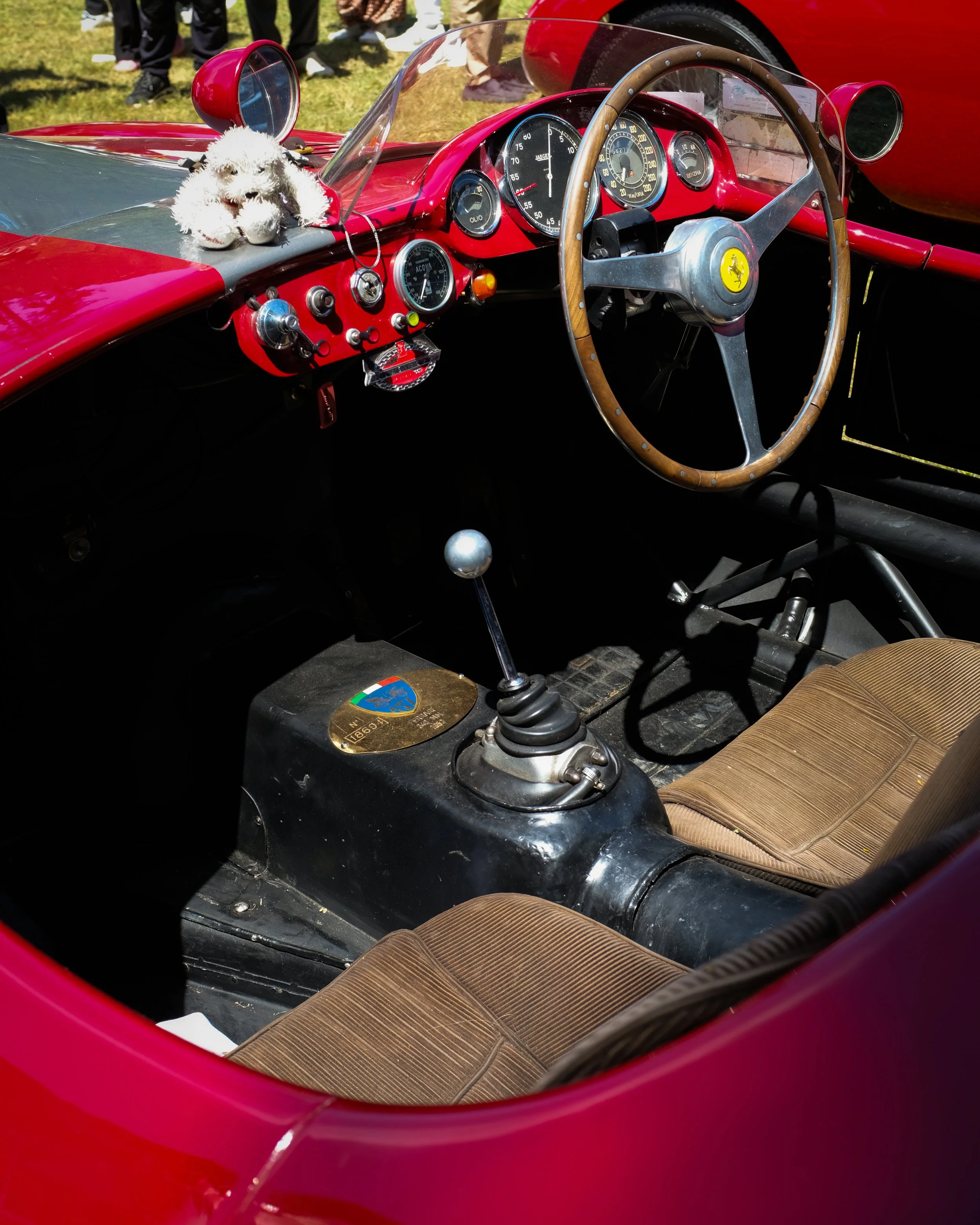 Interno di una vettura sportiva rossa Ferrari d'epoca, con volante di legno, strumenti analogici e sedili in tessuto marrone.