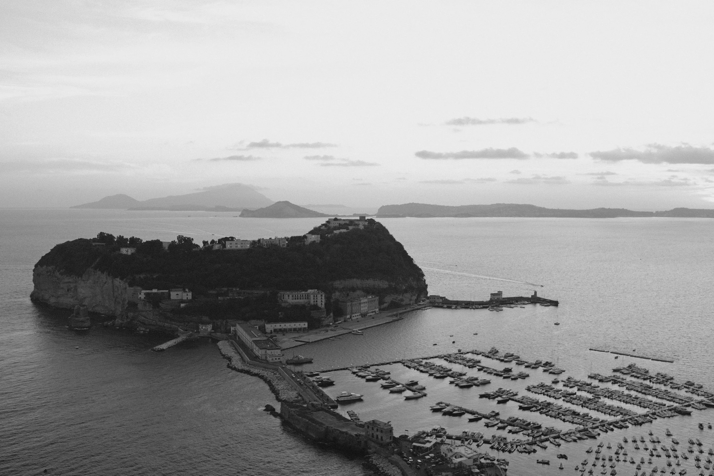 Veduta di un porto con molte barche ormeggiate, situato davanti a un'isola con alcune costruzioni e colline verdi, sul mare con isole in lontananza e cielo nuvoloso.