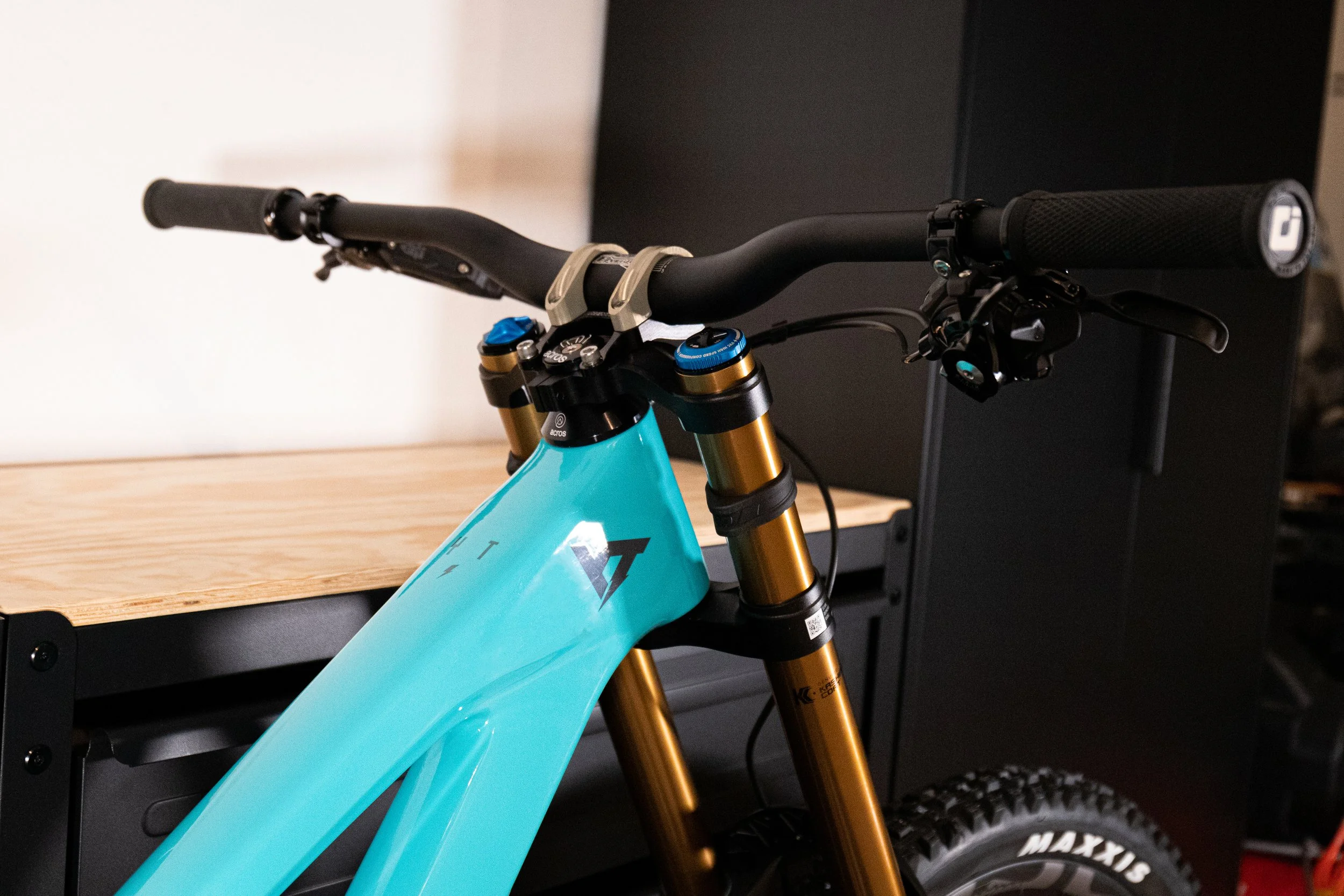 Bicicletta da downhill con telaio di colore azzurro, forcella anteriore dorata e manubrio nero, appoggiata a una superficie di legno e metallo in un ambiente interno.