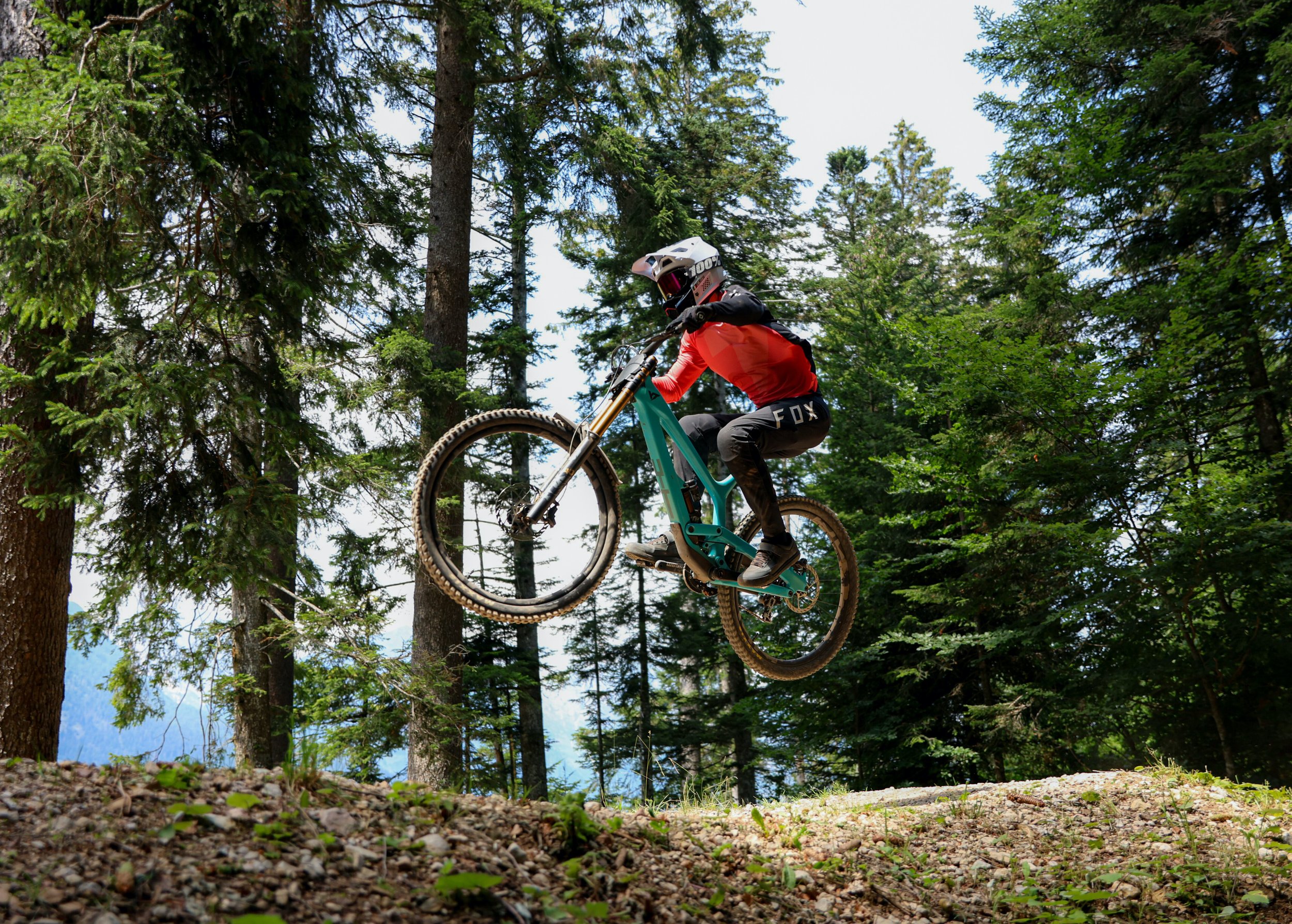 Un ciclista in abbigliamento da mtb, casco e visiera, salta sopra un terreno irregolare in una foresta di alberi alti e fogliame verde.