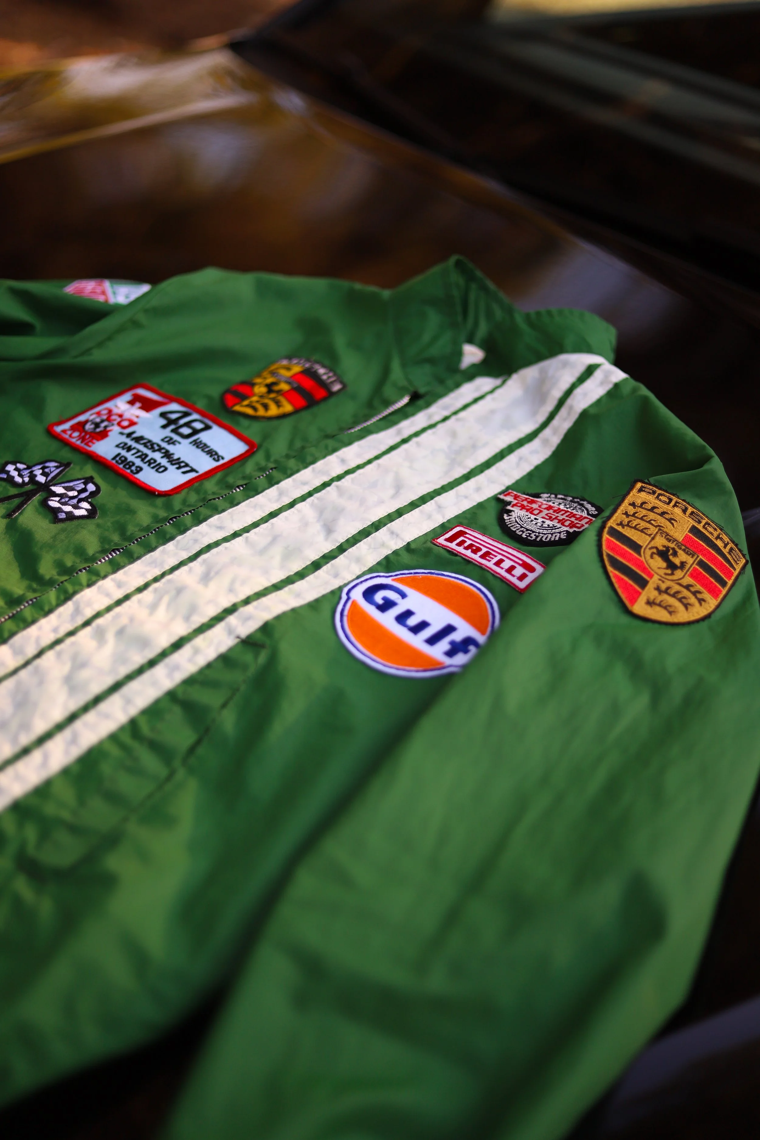 Giacca verde con patch di marchi automobilistici come Porsche, Gulf, Pirelli e altri, posata su una superficie di legno.