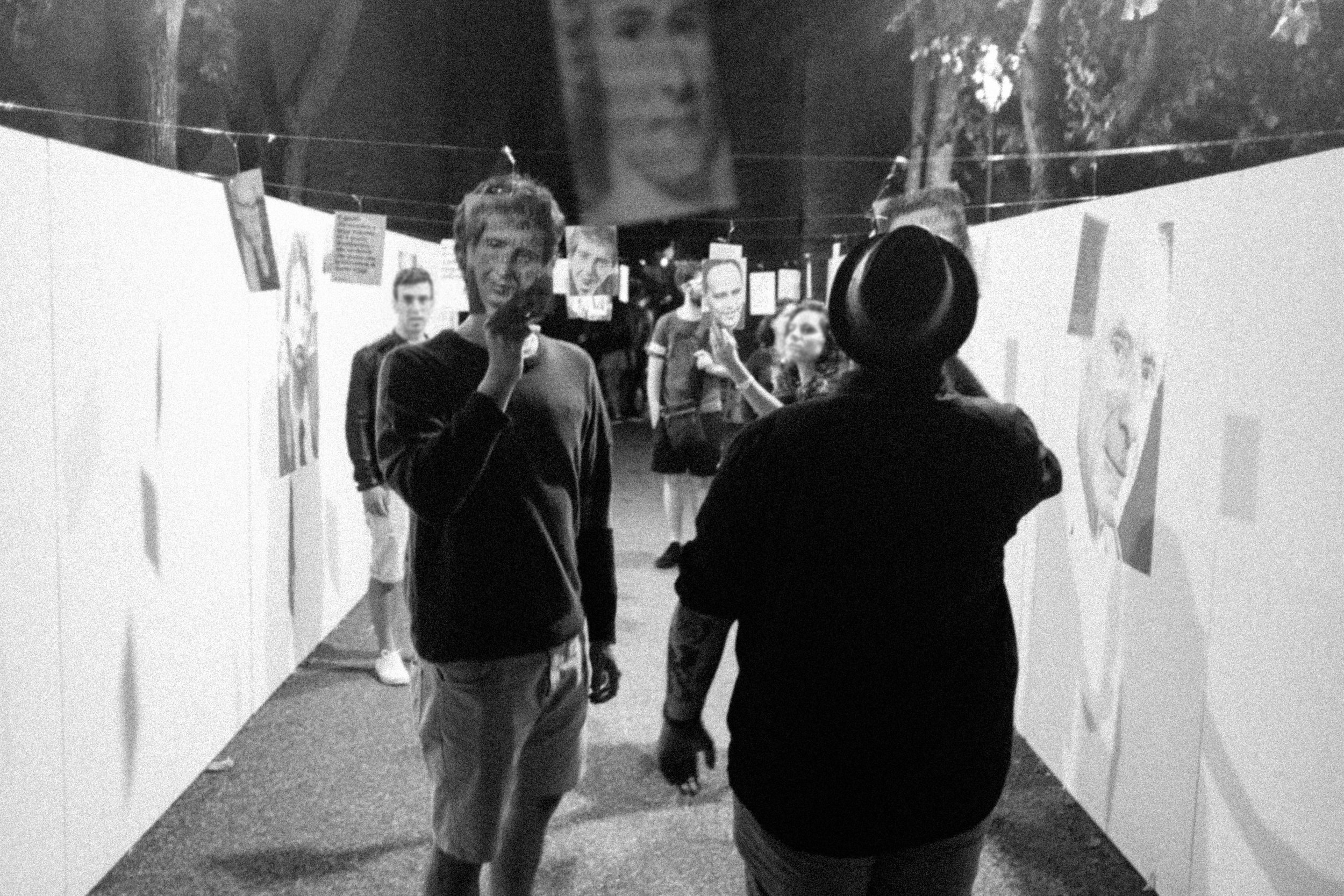 Galleria d'arte con persone che osservano e discutono opere d'arte, fotografie e disegni su pareti, in un ambiente illuminato.