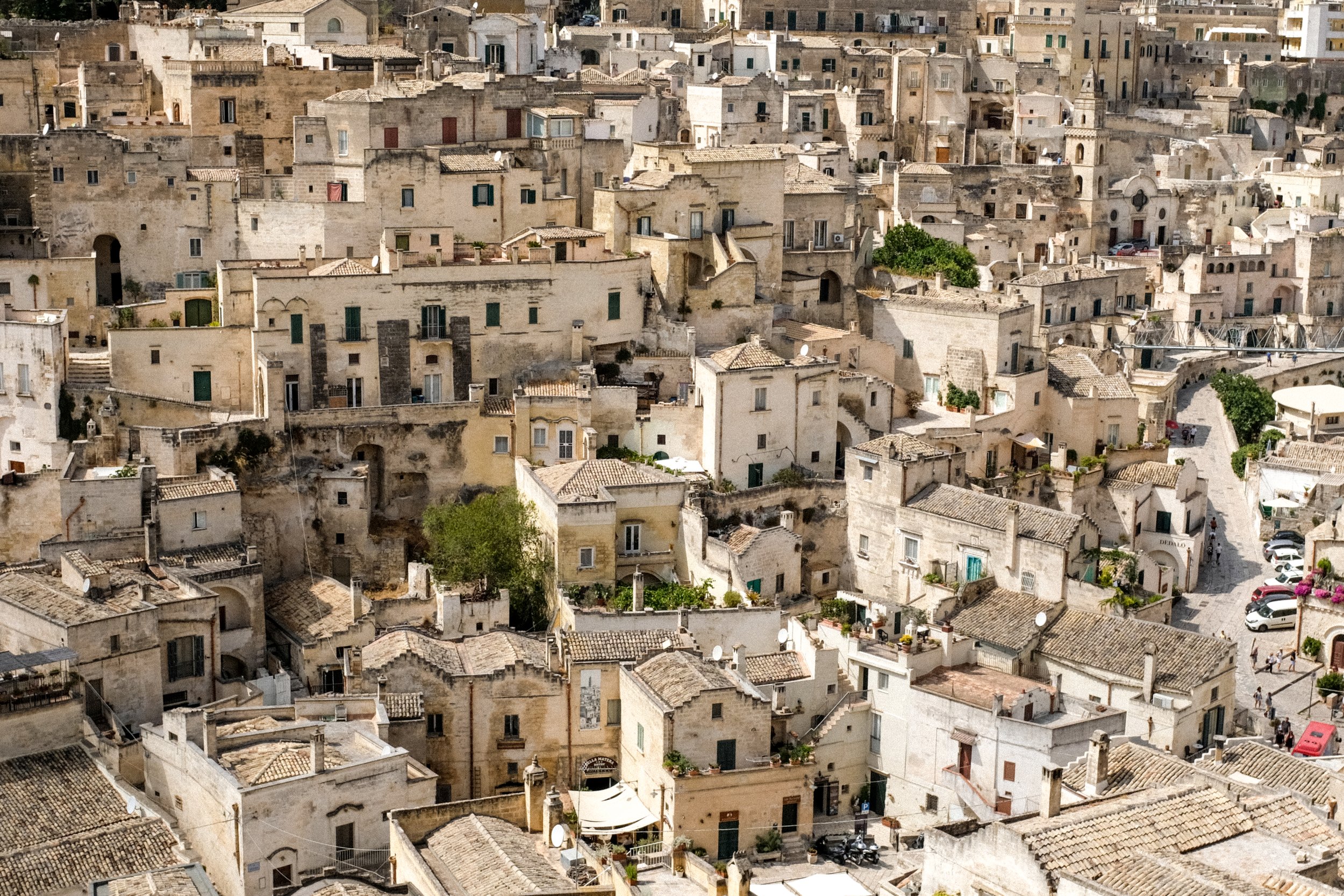 Veduta di un insieme di case di pietra con tetti in tegole, in un nel centro storico di una città italiana, con stretti vicoli, alcune macchine parcheggiate e piccole aree verdi.