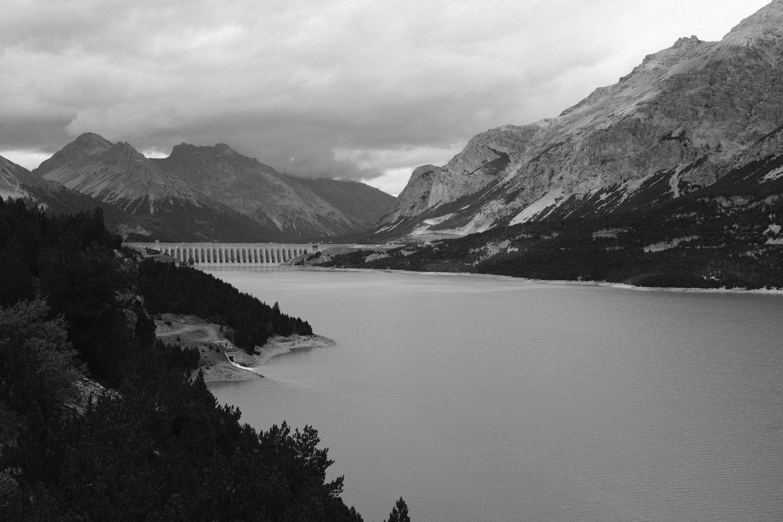 Paesaggio montano con un lago, una diga e montagne in lontananza, in bianco e nero.
