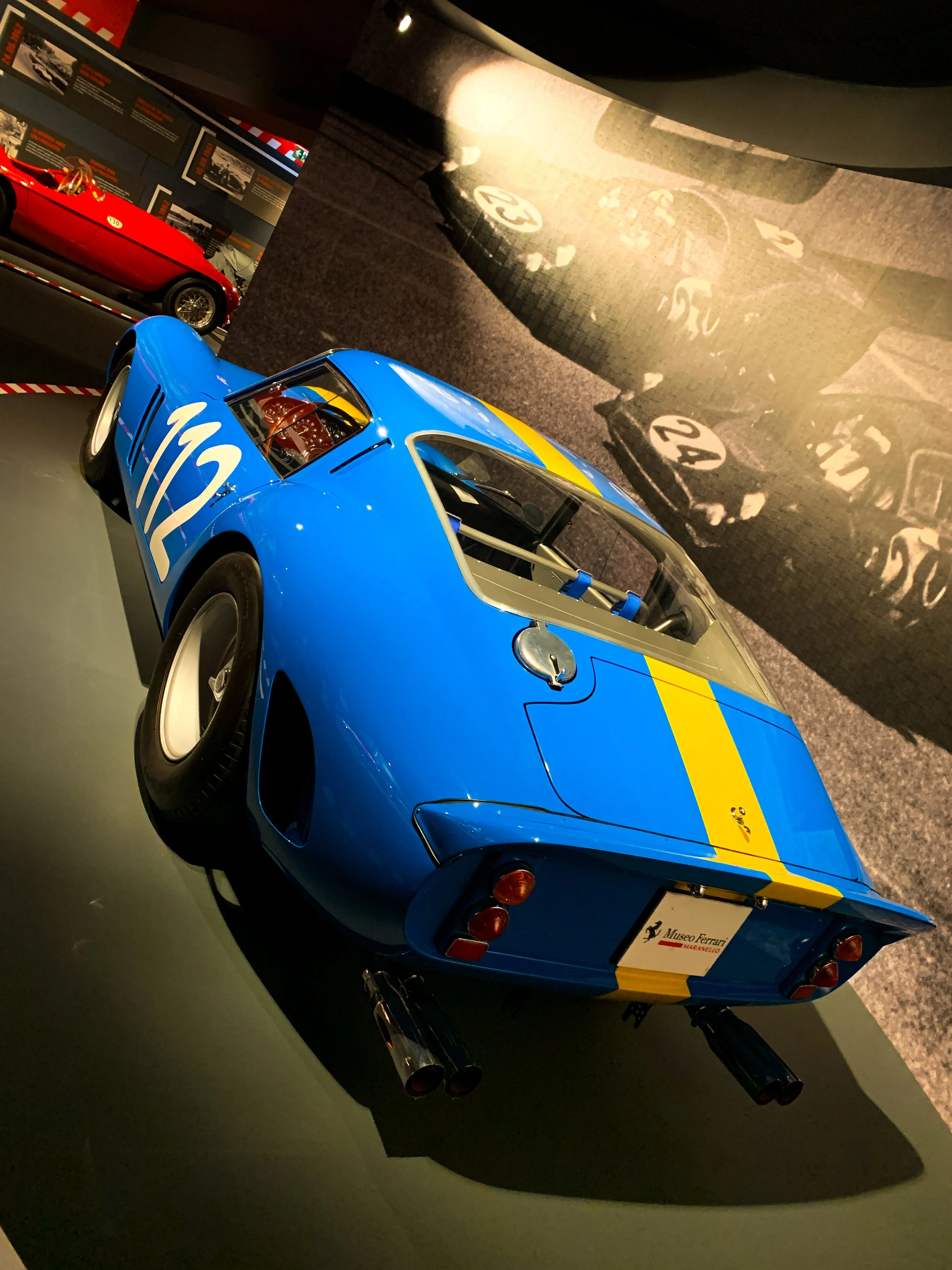 Auto da corsa Ferrari blu con strisce gialle, esposizione in un museo, con un cartellino identificativo e una grande foto storica sul muro di sfondo.