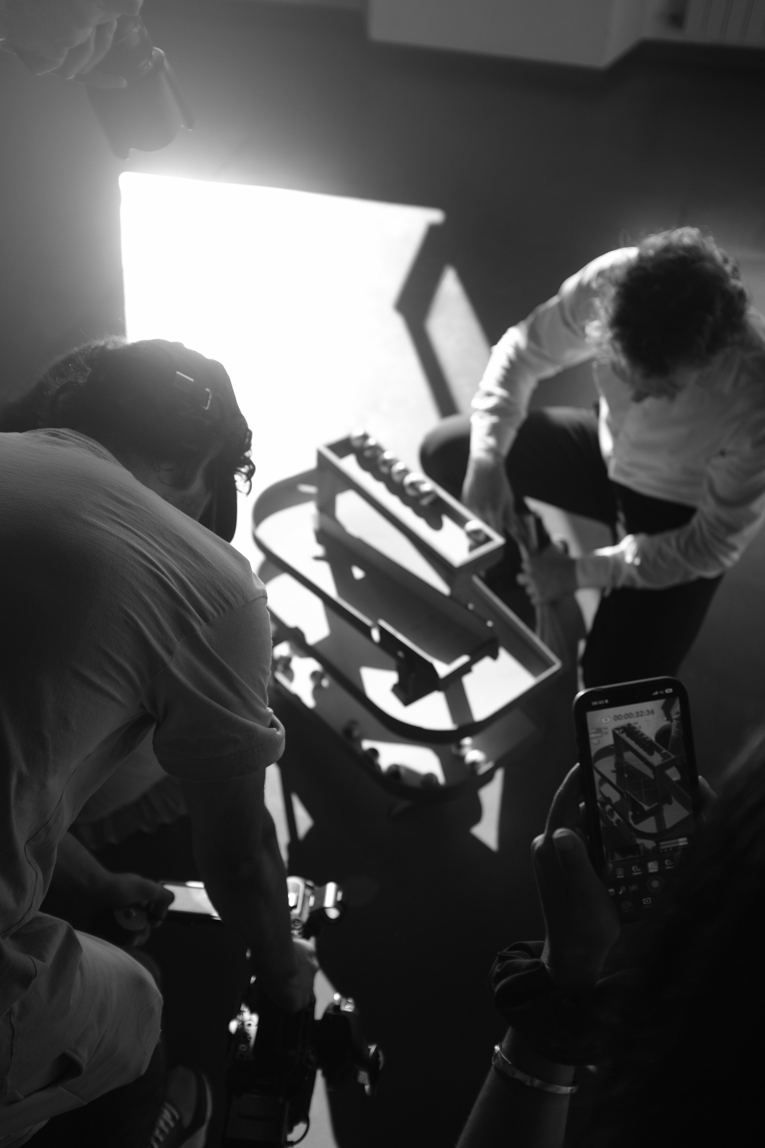 Tre persone sono intorno a una tavola da lavoro con un modellino di ferrovia, una fotocamera e uno smartphone. Una persona sta lavorando sul modellino, mentre un'altra registra la scena con il cellulare.