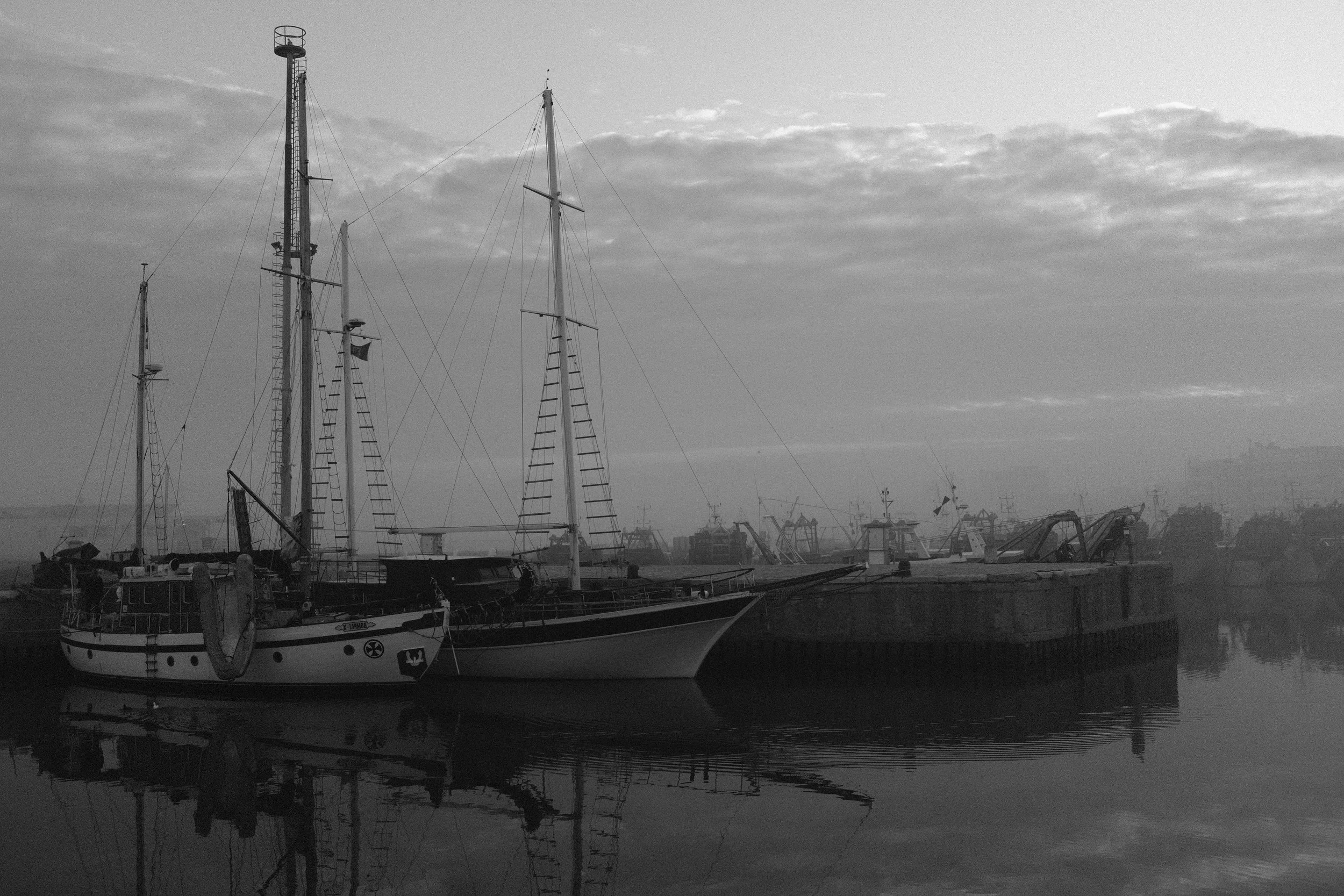 Foto di una zona portuale con barche a vela ormeggiate, in bianco e nero, con un cielo nuvoloso e un mare calmissimo