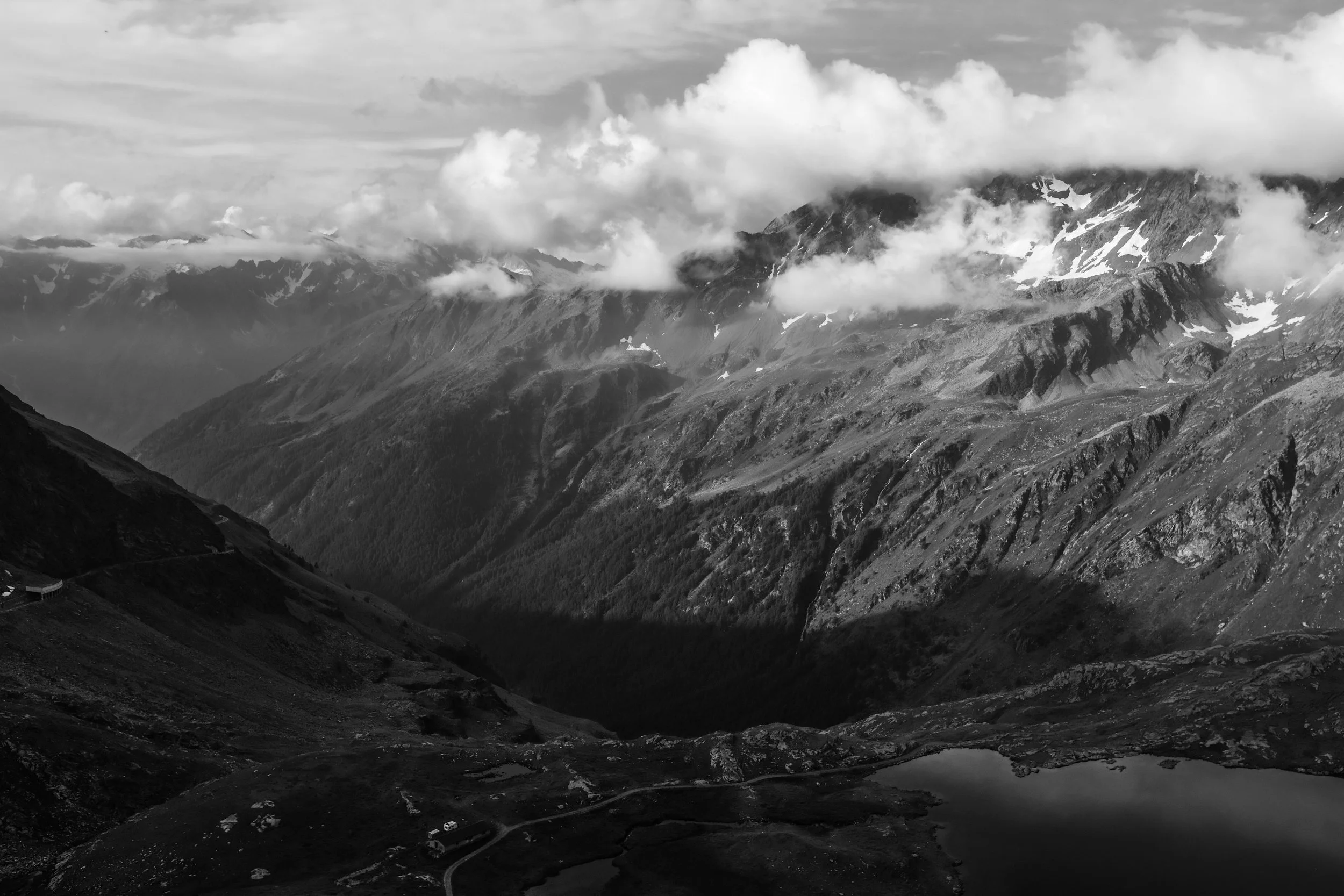 Paesaggio montano con nuvole, montagne con nevi temporanee, lago e strade nel primo piano, in bianco e nero.