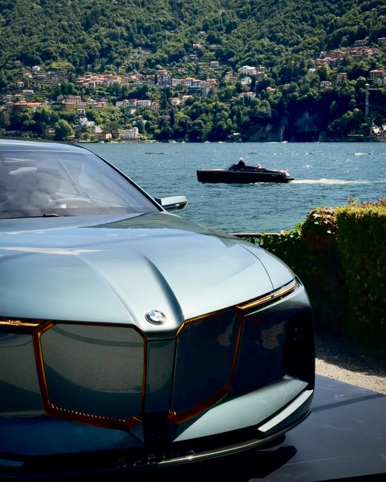 Parte anteriore di una macchina BMW di colore grigio scuro parcheggiata con un lago e una collina ricoperta di case e vegetazione sullo sfondo.