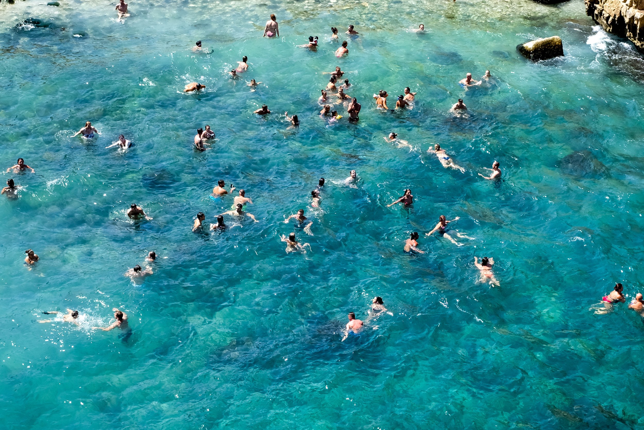 Persone che nuotano e si divertono nel mare cristallino con acque di diverse tonalità di blu, vicino a delle rocce.
