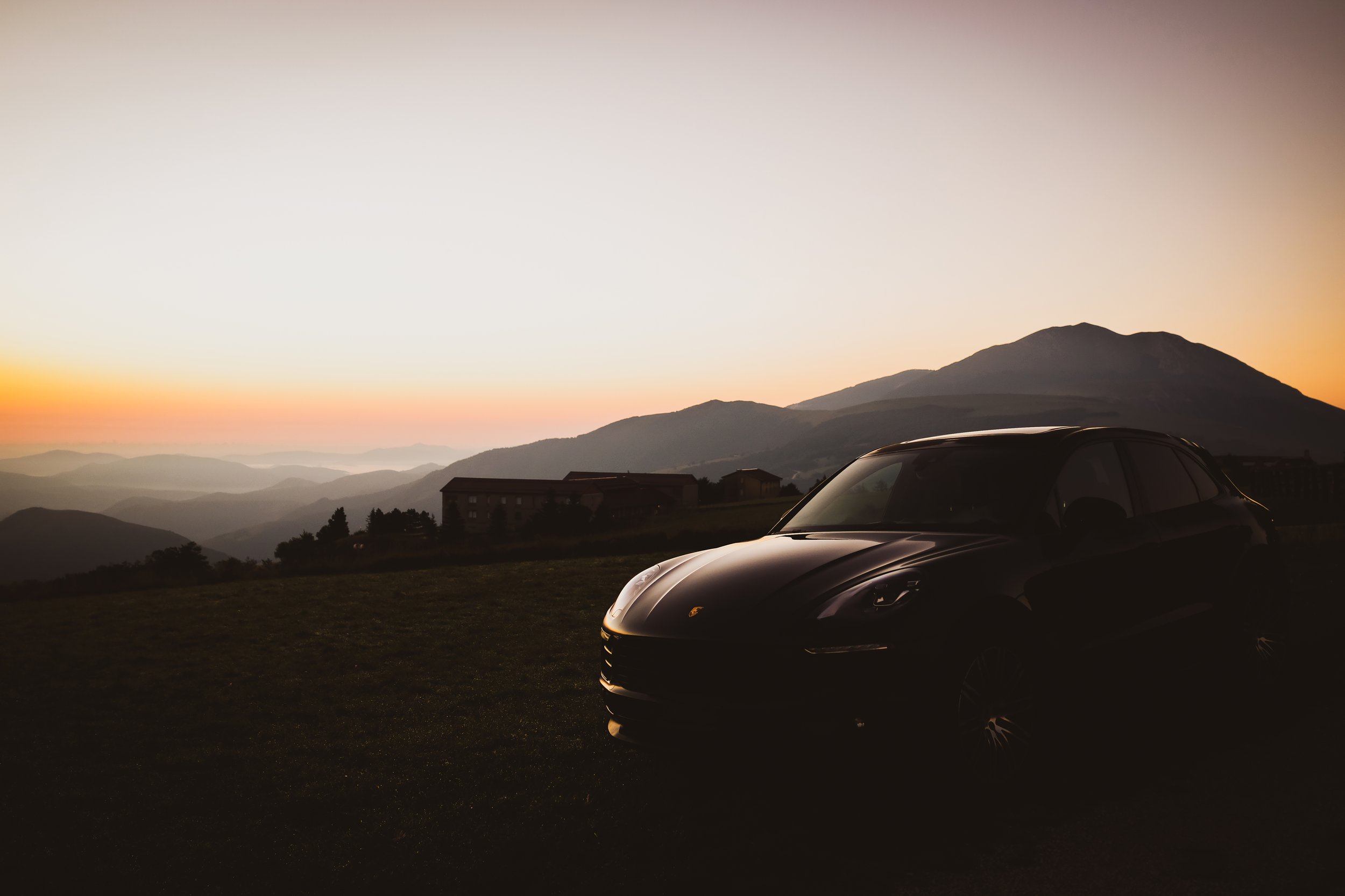 Auto sportiva nera in primo piano con paesaggio montano e tramonto sullo sfondo.