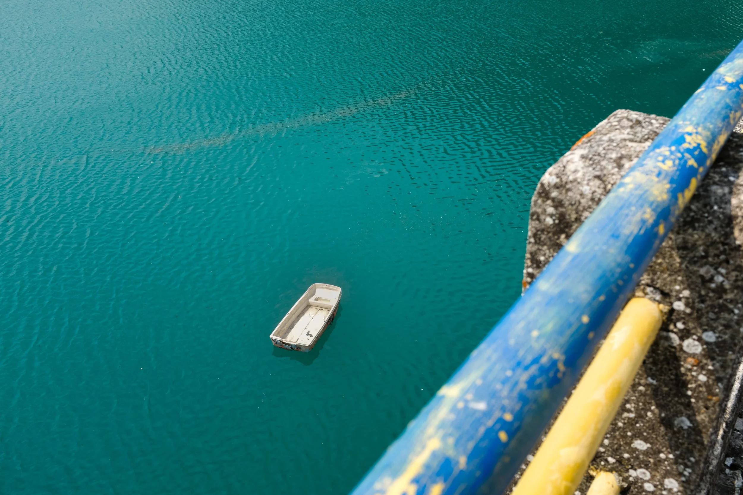 Barca abbandonata nel mare. Guardrail blu e giallo con graffiti, parete di pietra.