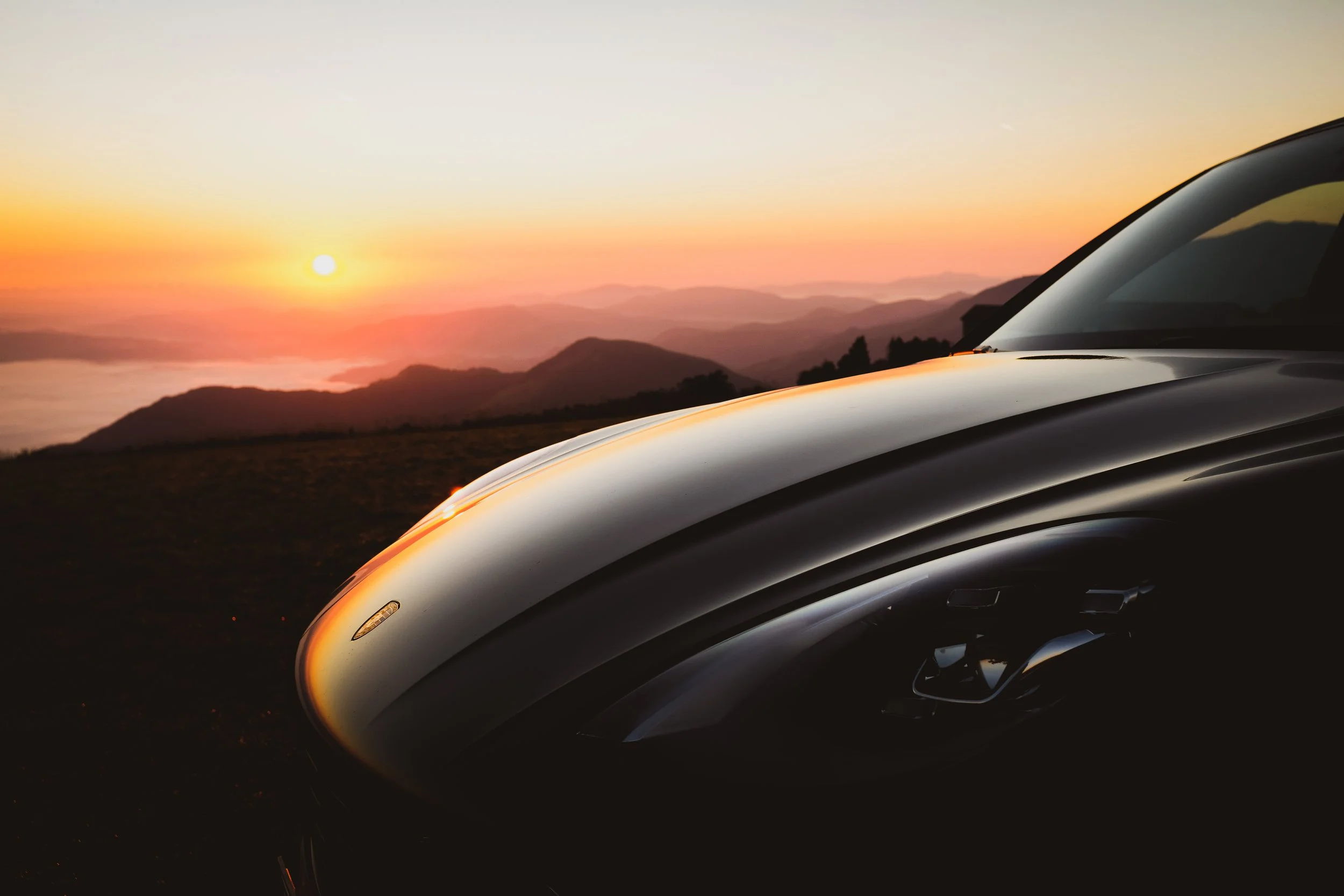 Particolare di un'auto nera davanti a un tramonto su una catena montuosa.