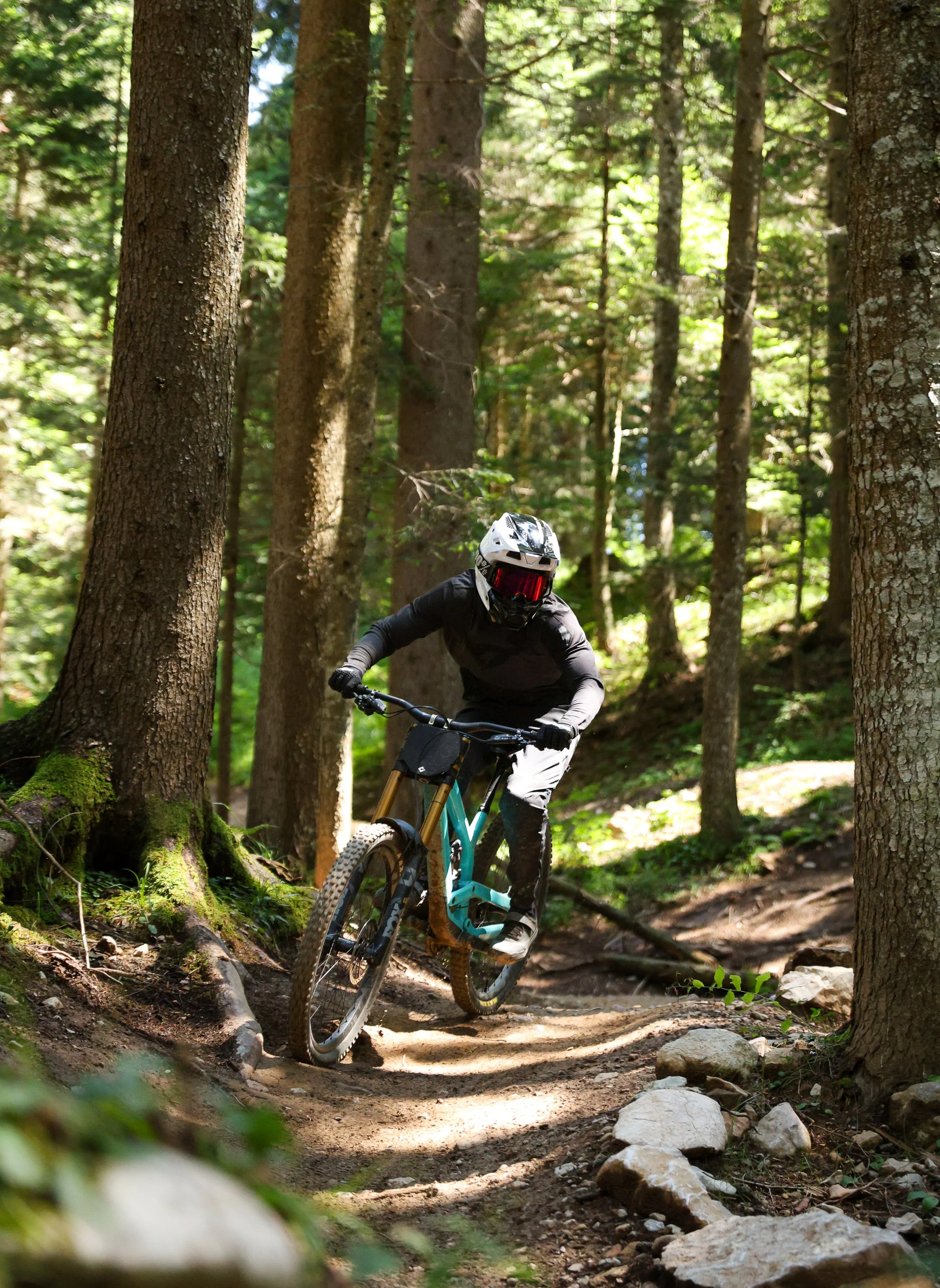 Un ciclista con casco e mascherina protettiva sta percorrendo un sentiero nel bosco con alberi alti e foglie verdi, in un'escursione di mountain biking.