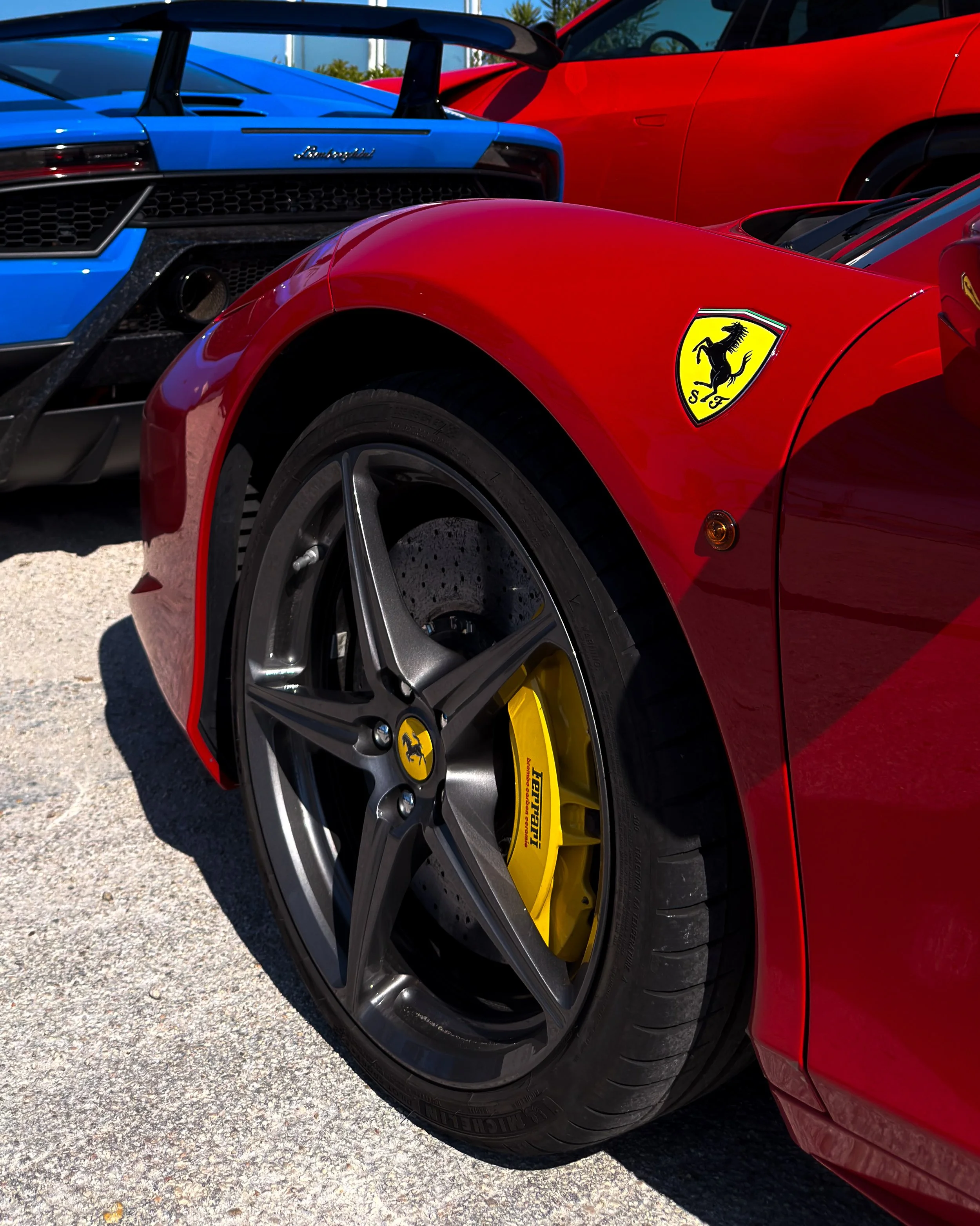 Foto di tre auto sportive di alta gamma, tra cui una Ferrari rossa, una Lamborghini blu e una seconda auto rossa. La macchina Ferrari ha i cerchi sportivi con il logo Ferrari e un adesivo con il nome Pirelli.