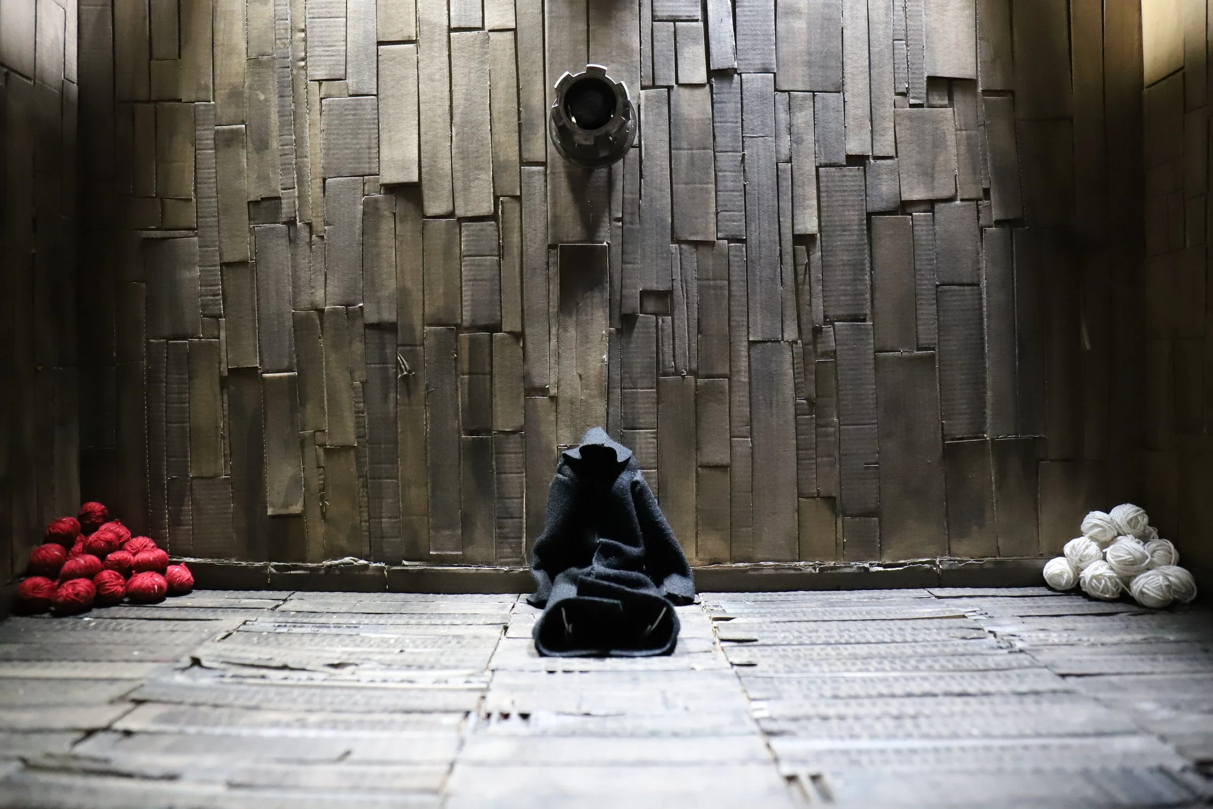 Una stanza con pareti e pavimenti in legno, con palle di filo di lana rosse sulla sinistra e bianche sulla destra, e un cappotto nero appoggiato a terra di fronte a una parete.