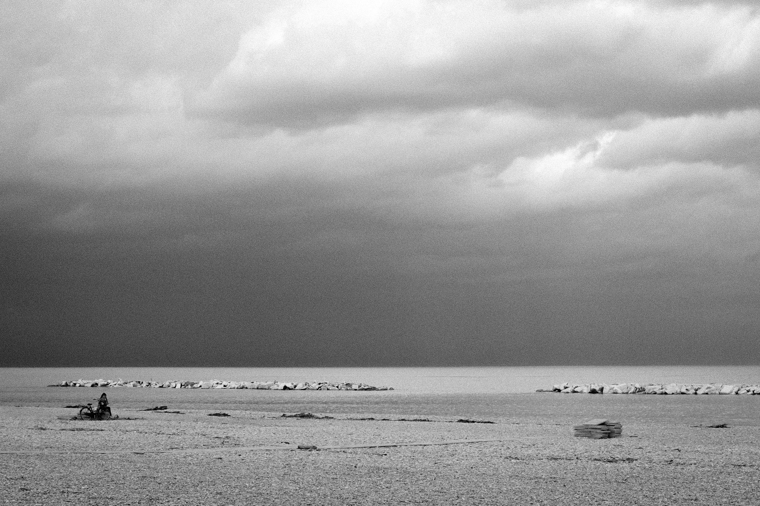 Spiaggia vuota con alcune rocce e una bicicletta distesa sulla sabbia, cielo nuvoloso in bianco e nero.