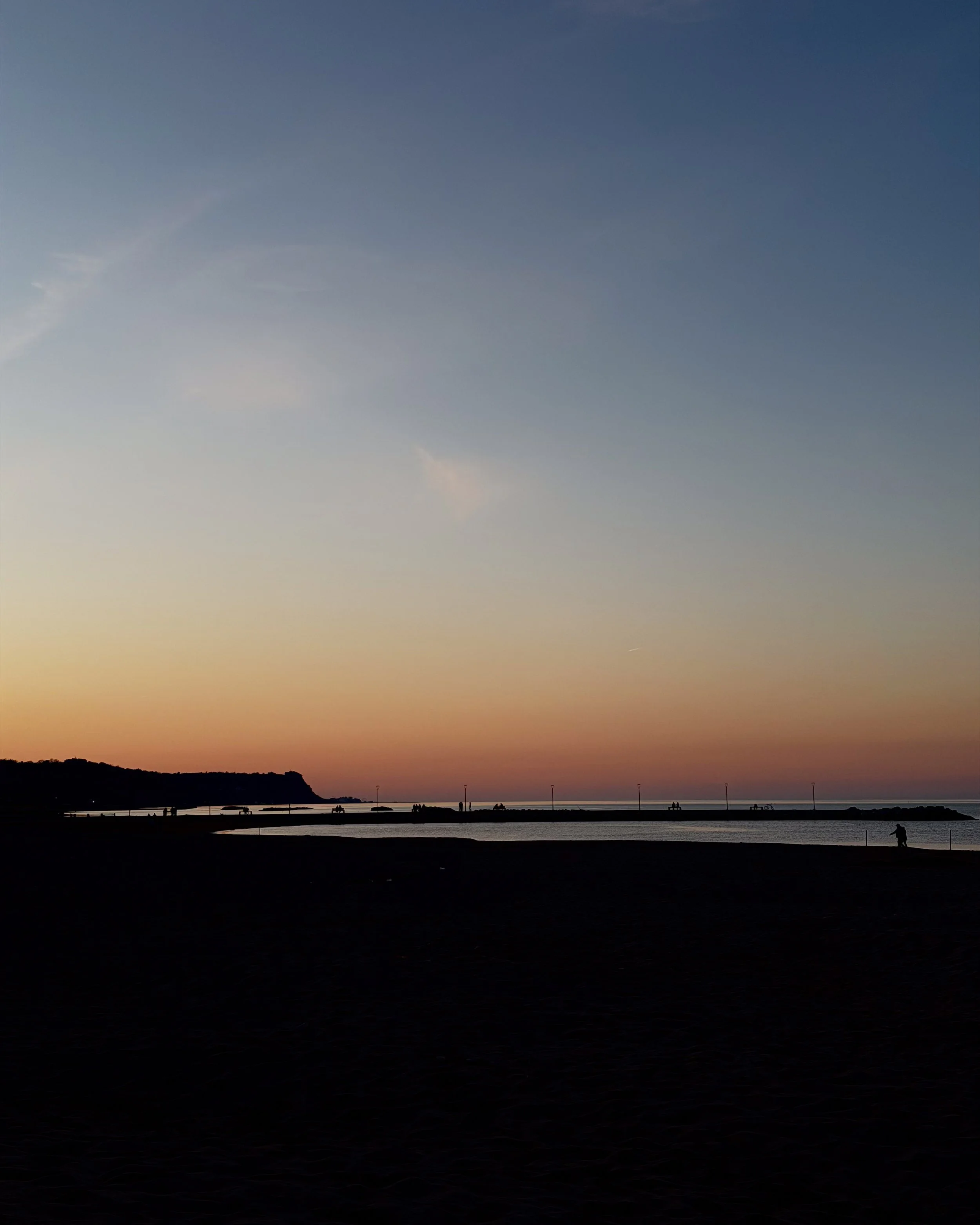 Tramonto sulla spiaggia con silhouette di una persona che cammina e barche in lontananza.