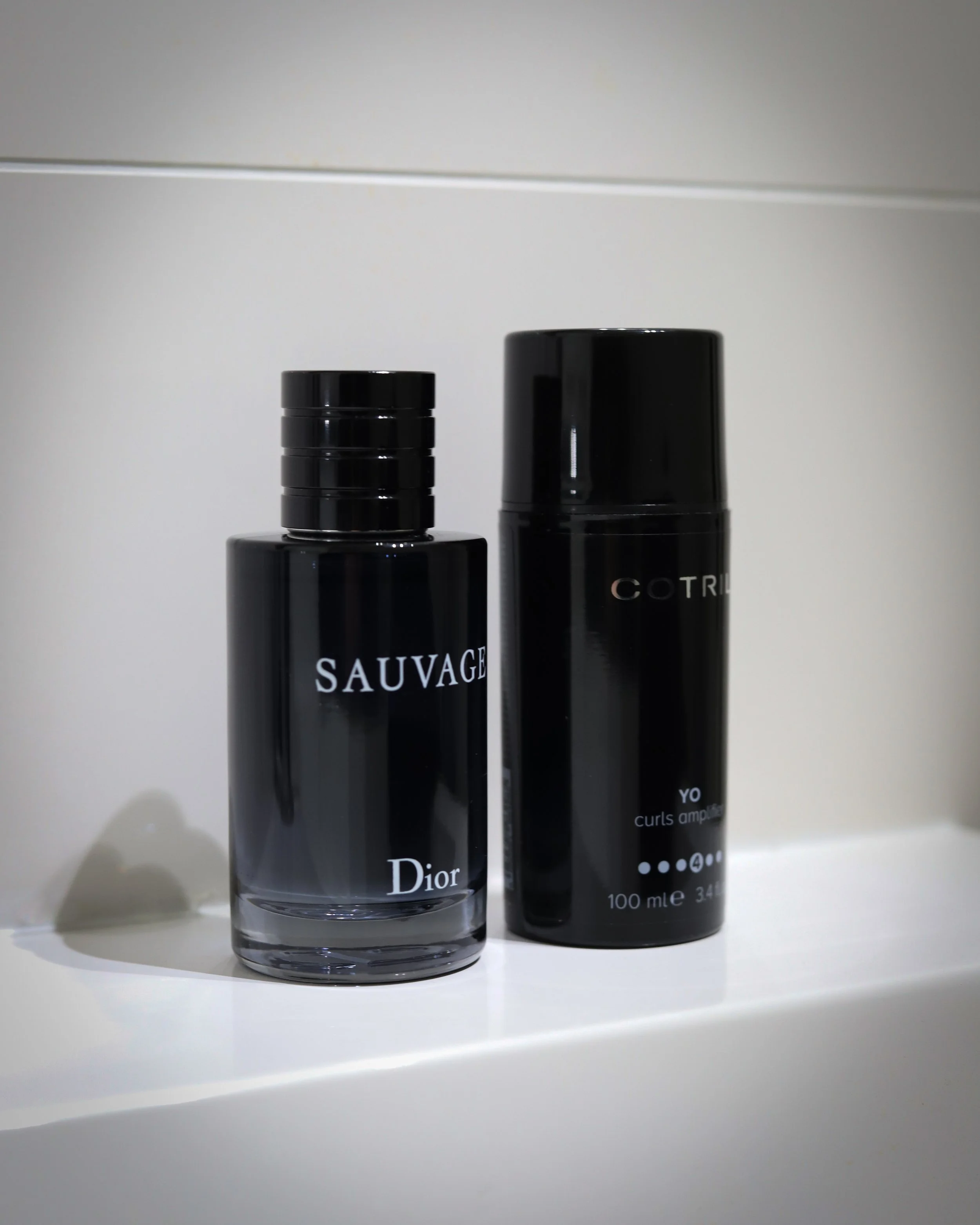 Due prodotti di cosmetici neri in bottiglie di plastica. La bottiglia a sinistra ha etichetta con scritto "SAUVAGE" e "Dior". La bottiglia a destra ha etichetta con scritto "COTR" e "yo curls amplified".