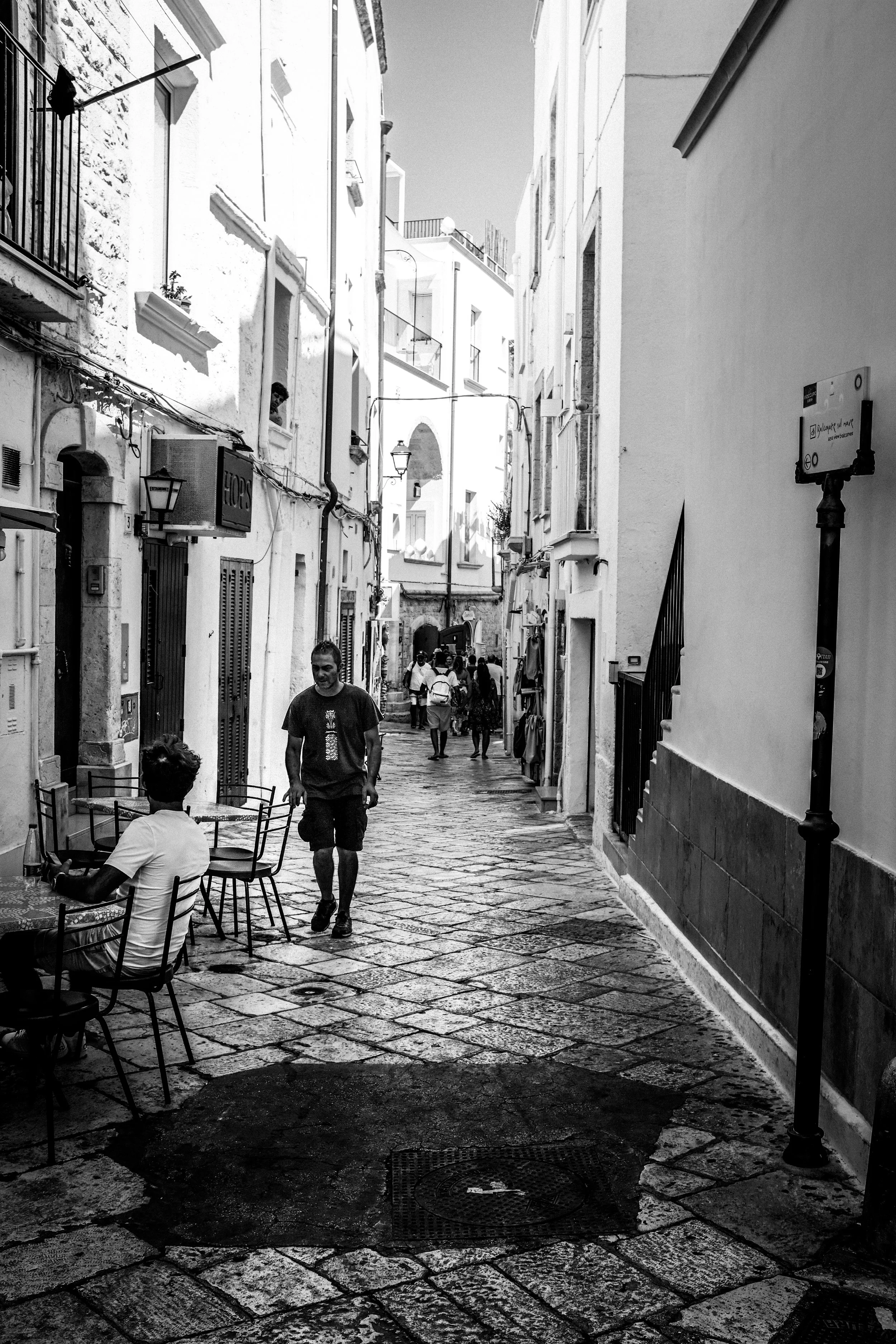 Via stretta e pavimentata in pietra, con edifici bianchi ai lati, alcune persone camminano e altre sono sedute ai tavoli di un bar; l'atmosfera è urbana e vivace.