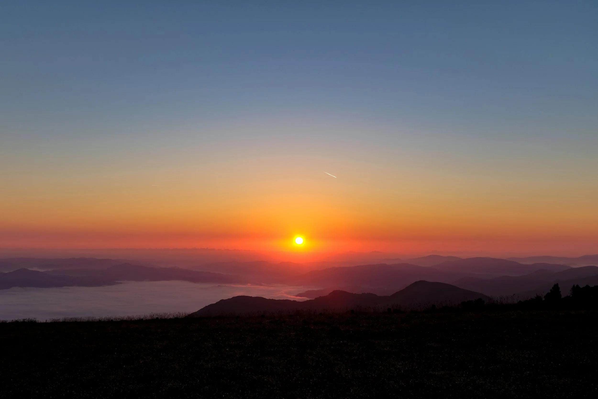 Sole che tramonta dietro le montagne al mattino, cielo colorato con sfumature arancioni, rosa e blu, con una foschia sulla valle sottostante.