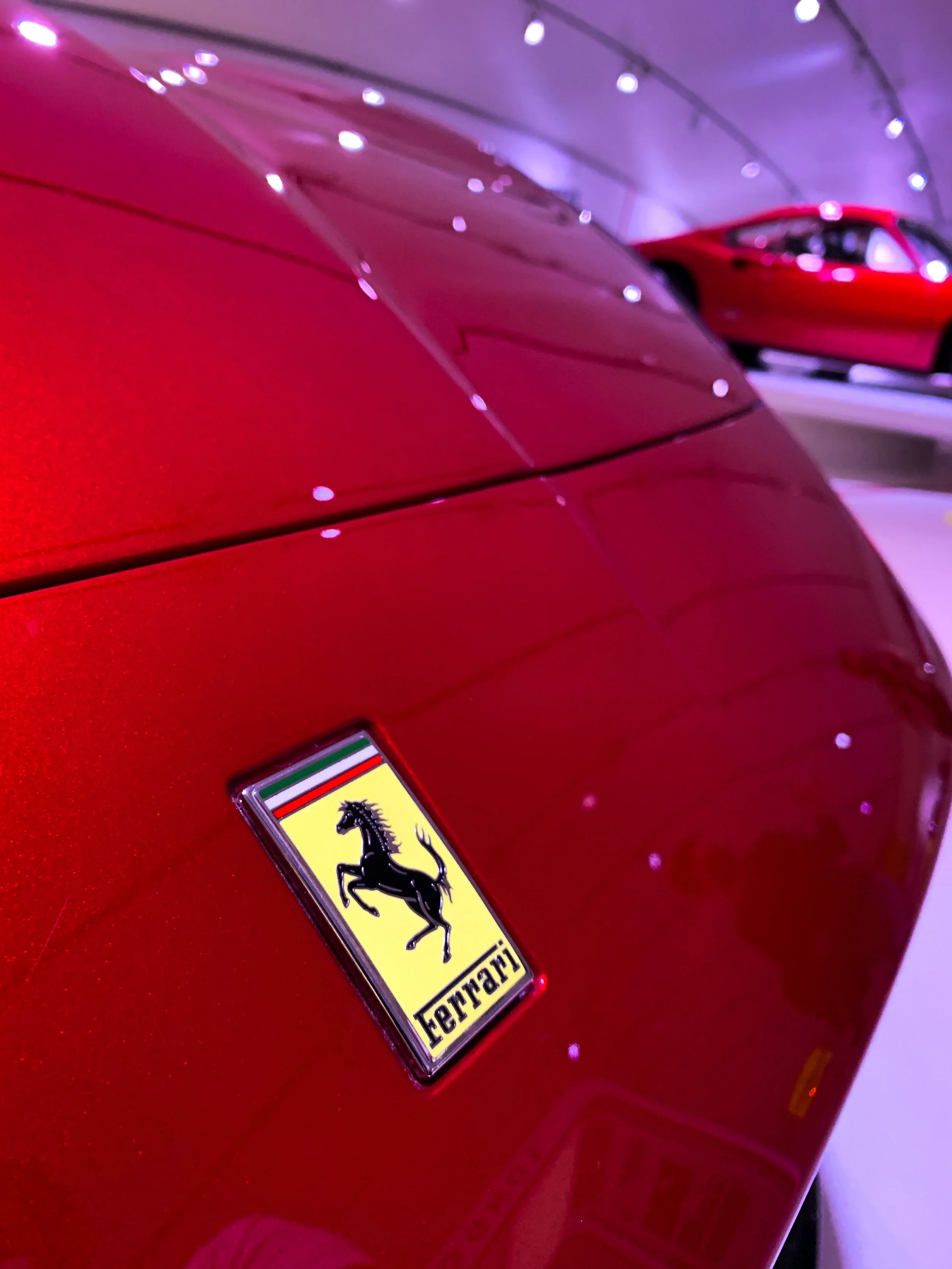 Primo piano di un'auto sportiva rossa Ferrari con il logo Ferrari visibile e un'altra auto rossa in background, in un ambiente lucido e ben illuminato.