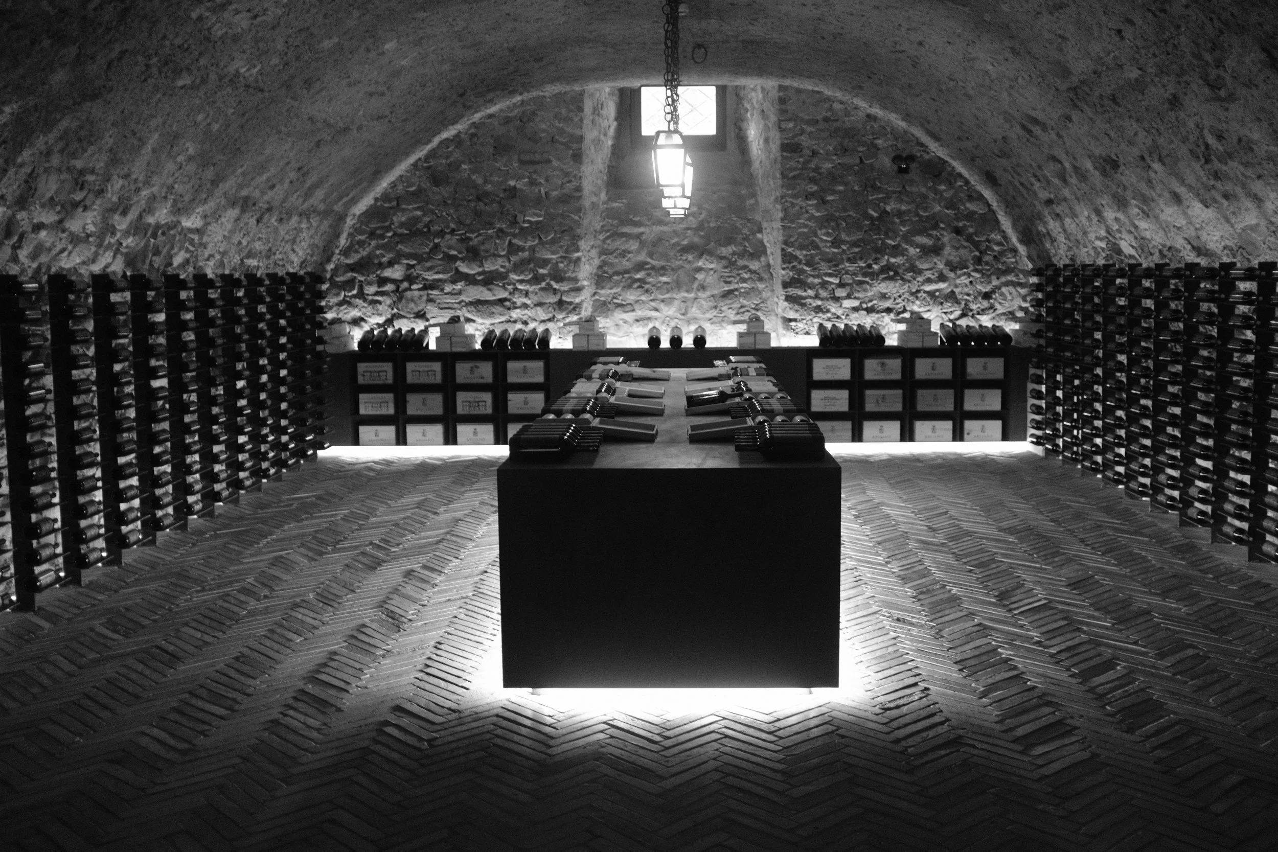 Botte di vino stoccate in una cantina sotterranea con pareti di pietra, tavolo centrale con bottiglie e bicchieri, illuminazione soffusa.
