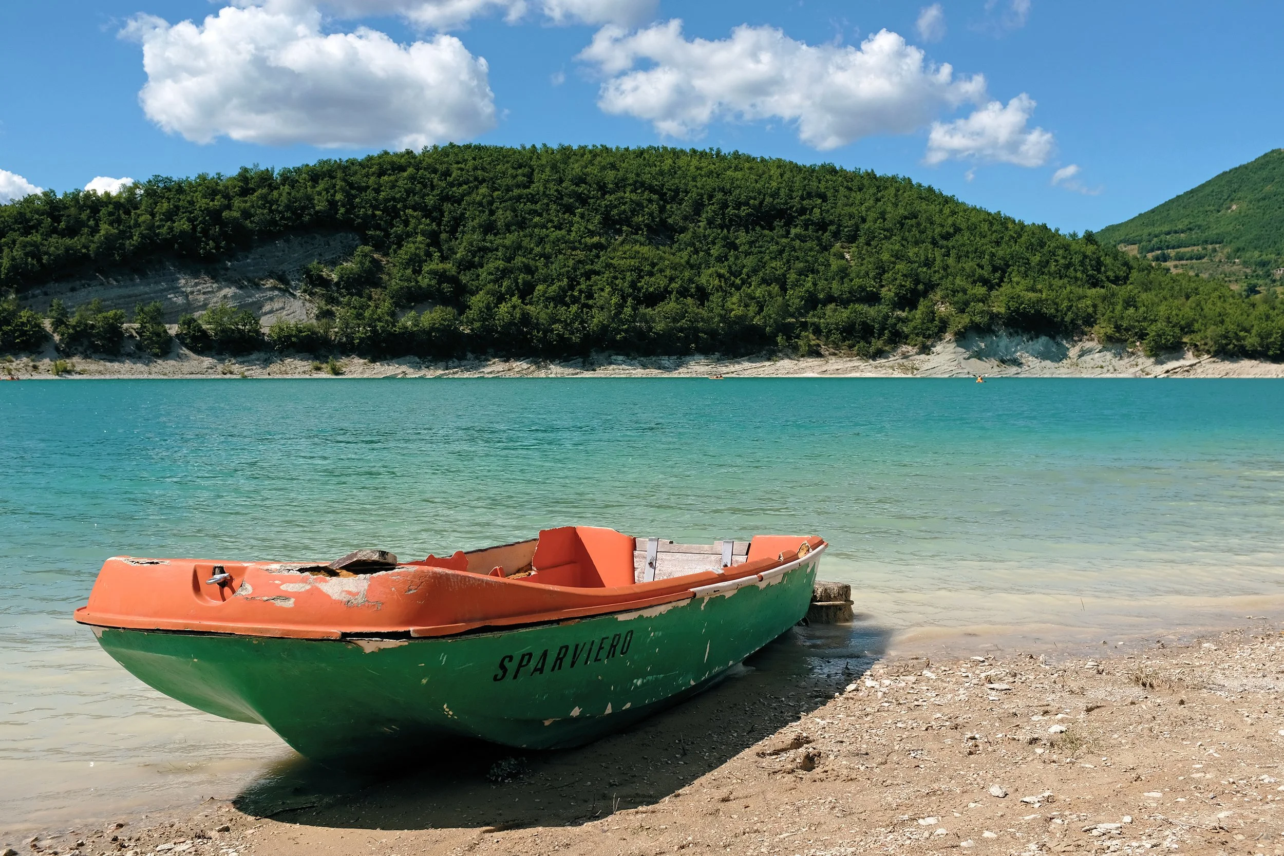 Barca abbandonata sulla spiaggia di un lago o mare, con acque blu e uno sfondo di colline verdi e un cielo con nuvole