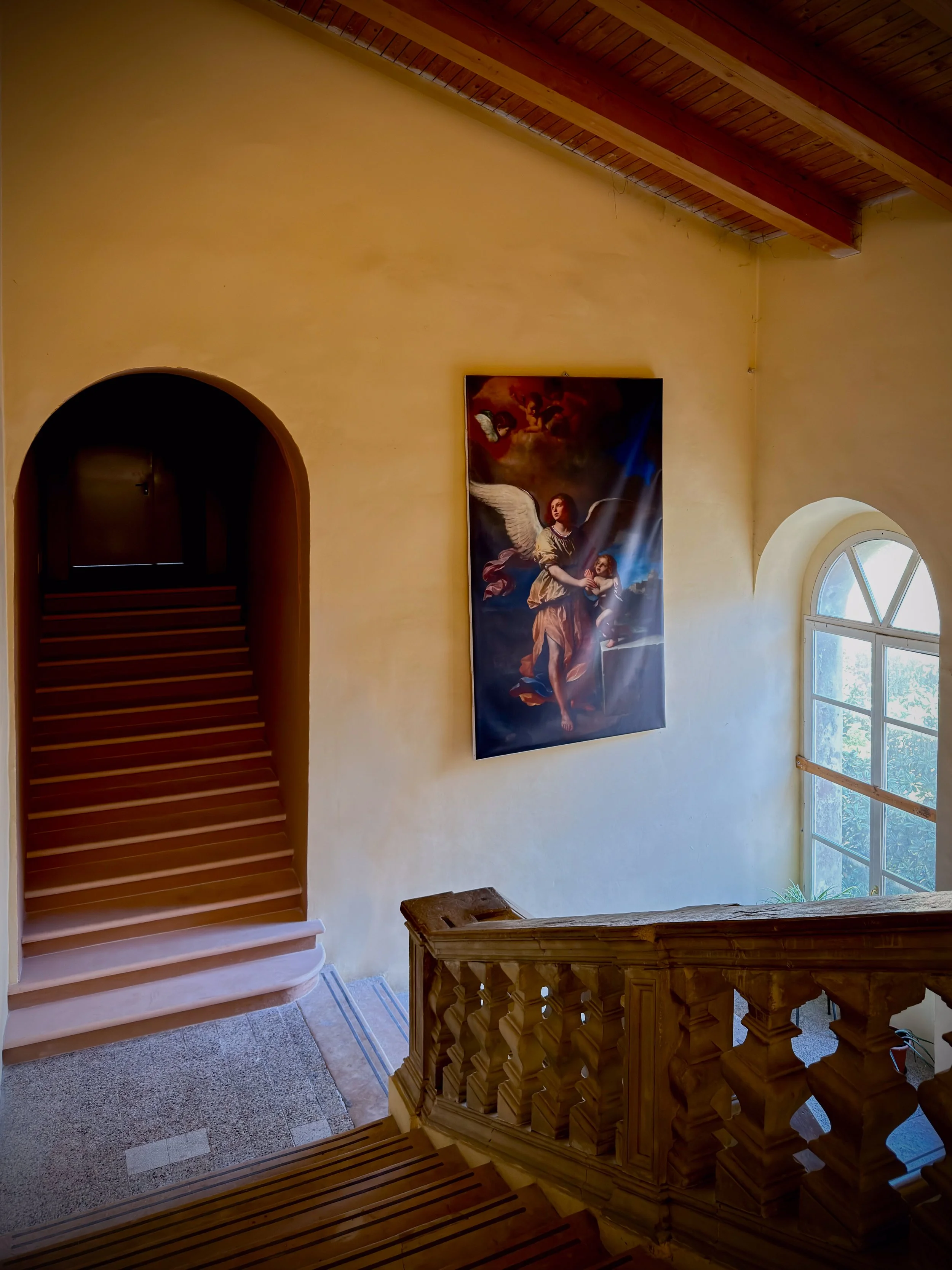 Interno di una casa con una scala in legno, una finestra e un dipinto sacro raffigurante un angelo con una spada.