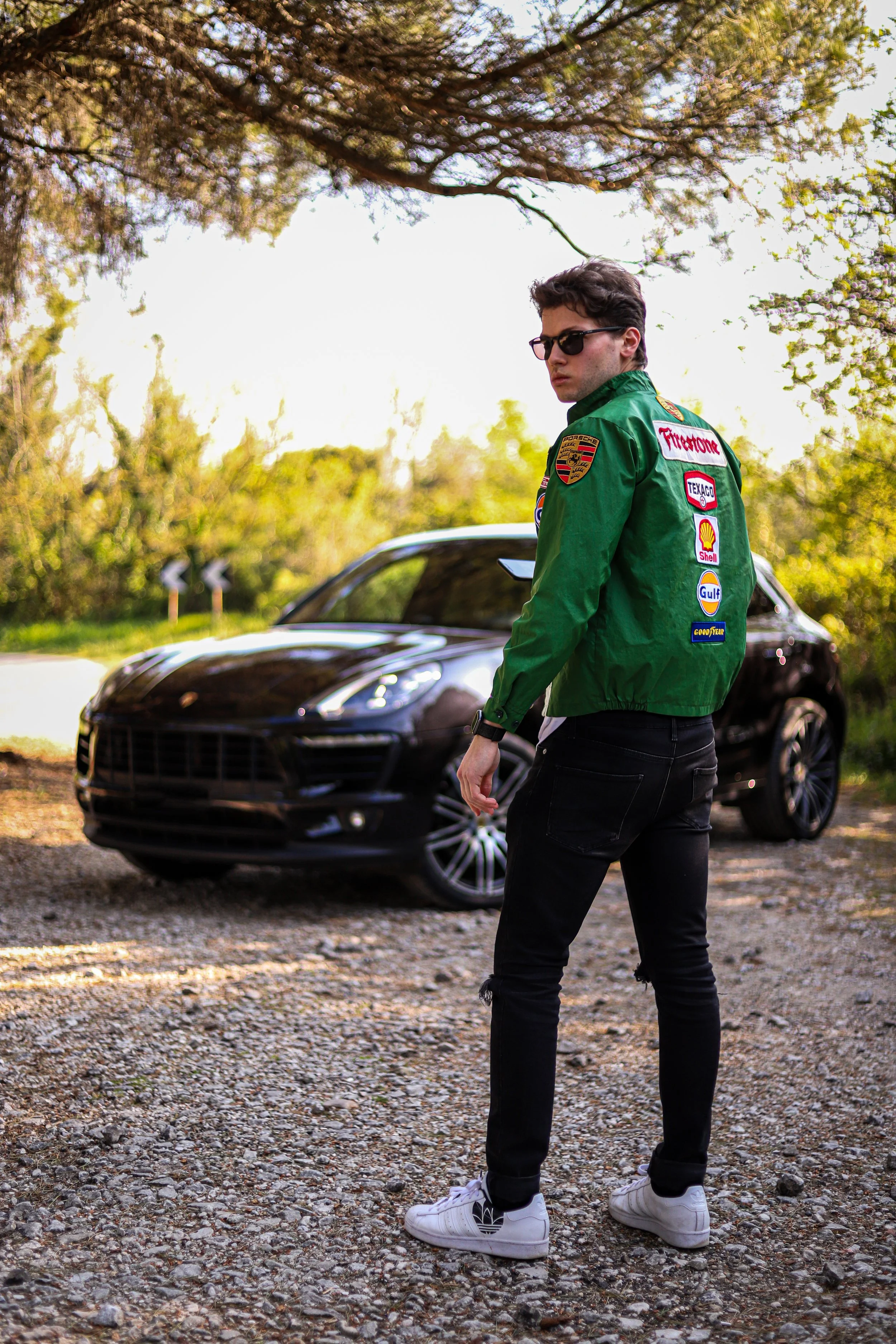 Un giovane uomo con occhiali da sole e giacca con patch di marchi automobilistici davanti a una macchina nera su un percorso sterrato in un'area verde.