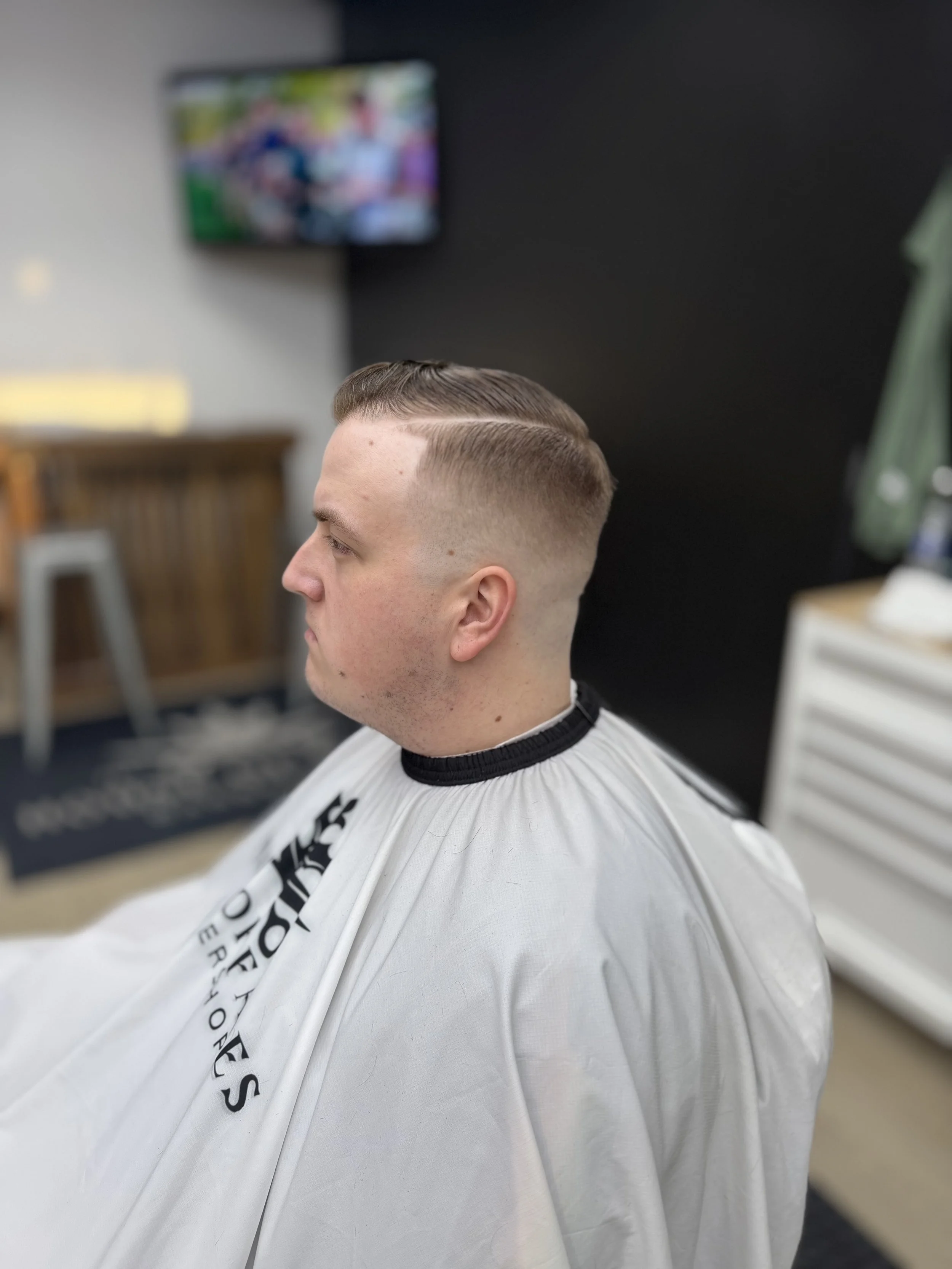 Side view of a man with a freshly cut, styled haircut, sitting in a barber's chair with a cape. The background shows a TV screen and barber shop interior.