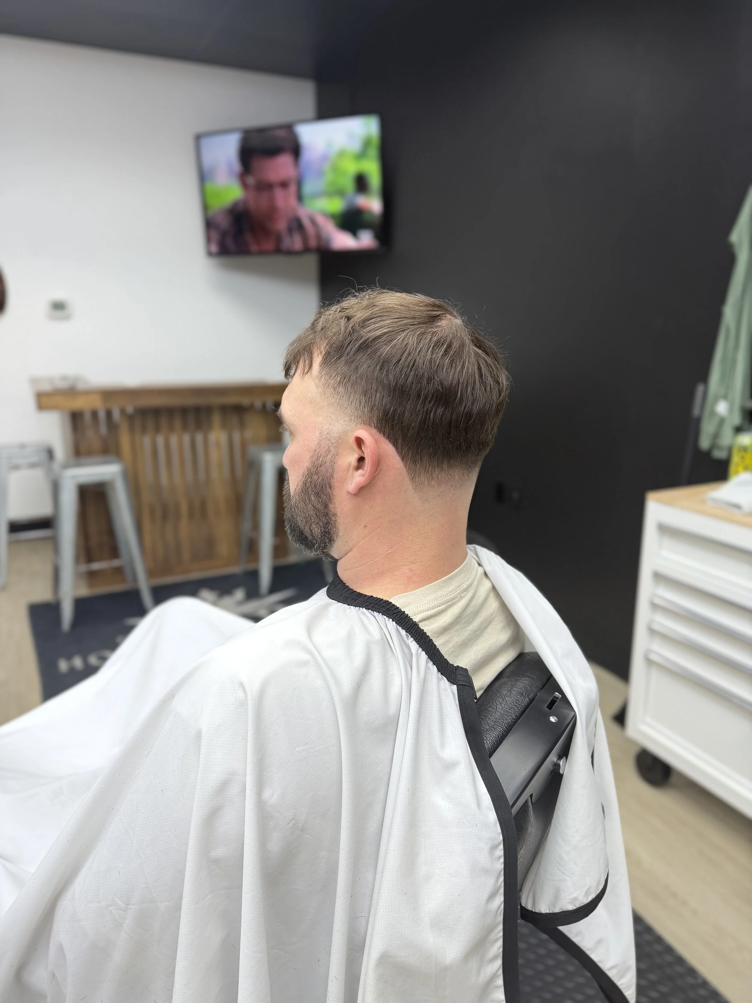 Man with freshly cut hairstyle sitting in a barber's chair, facing away from the camera, watching television in a barbershop.