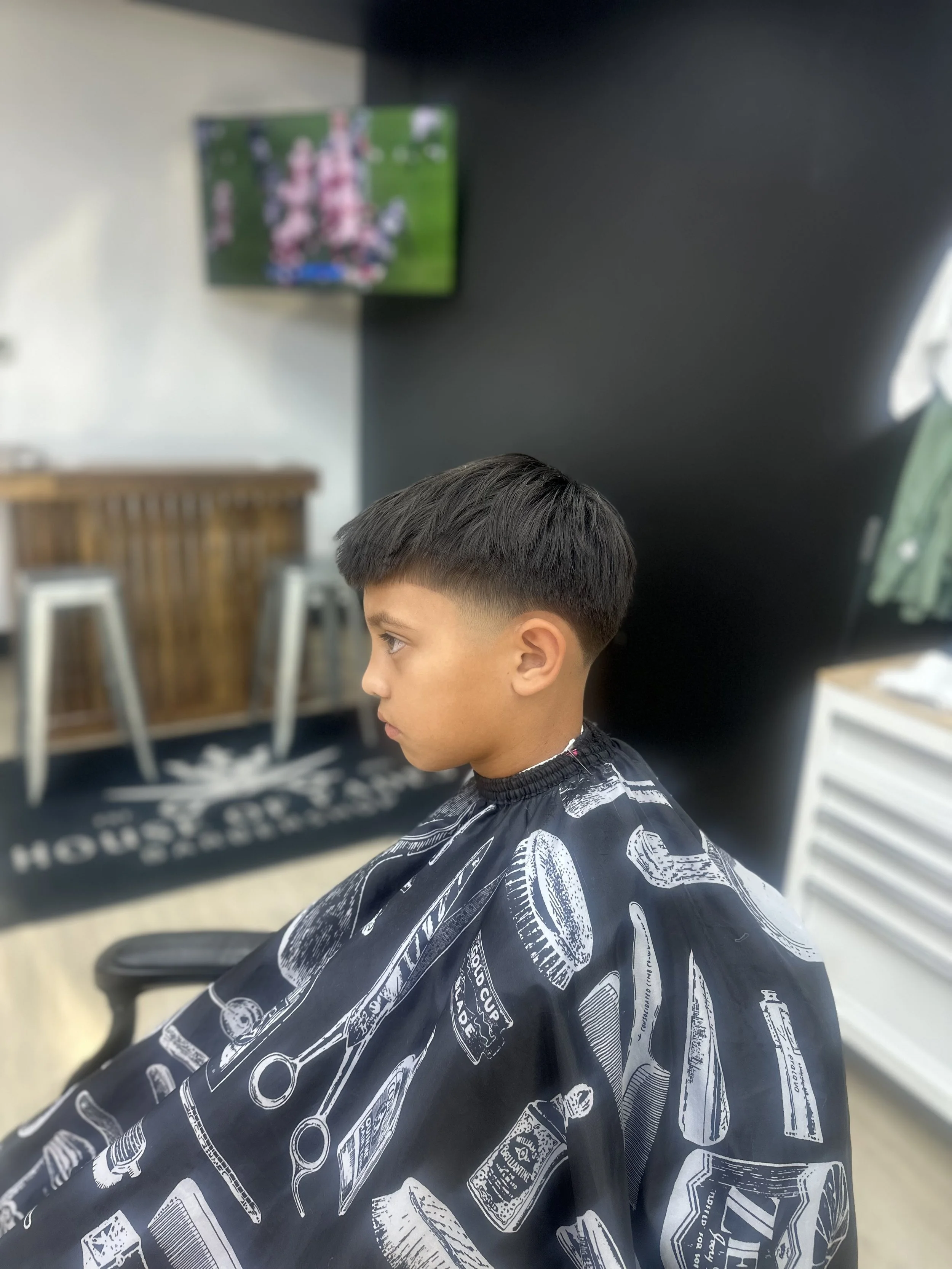 A young boy with a fresh haircut sitting in a barbershop, wearing a black cape with white barber-themed illustrations, watching a TV on the wall.