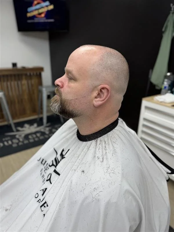 A man with a shaved head and beard sitting in a barber's chair, wearing a barber cape, in a barbershop.