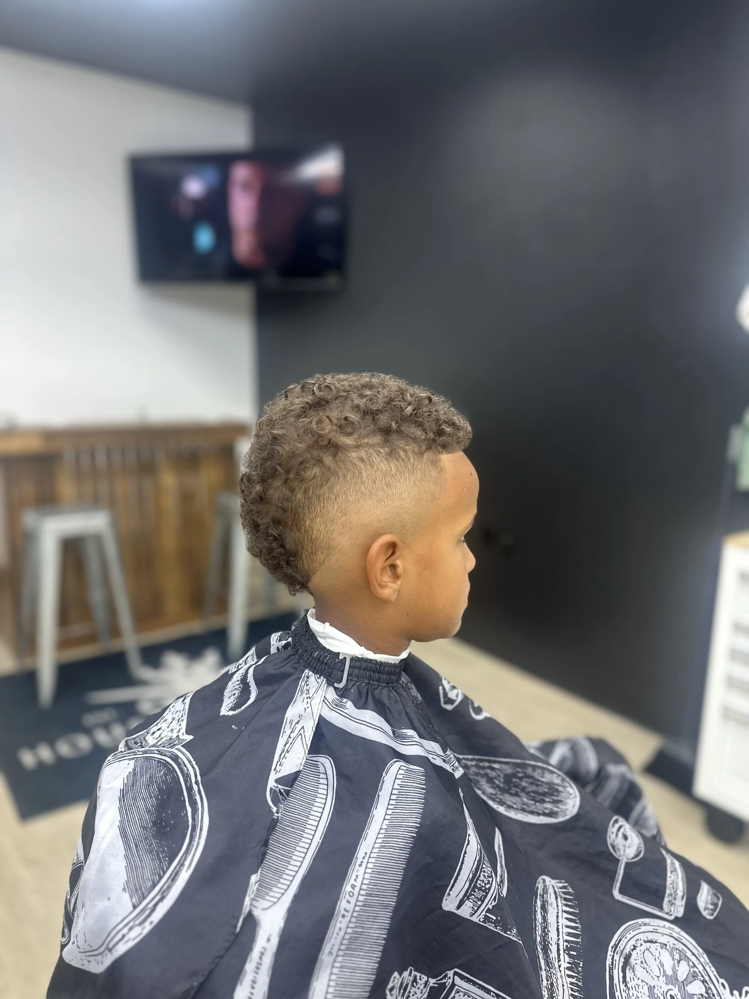 A boy has a new haircut in a barbershop, sitting in a black barber cape with barber tools illustrated on it, in a modern salon with a television in the background.
