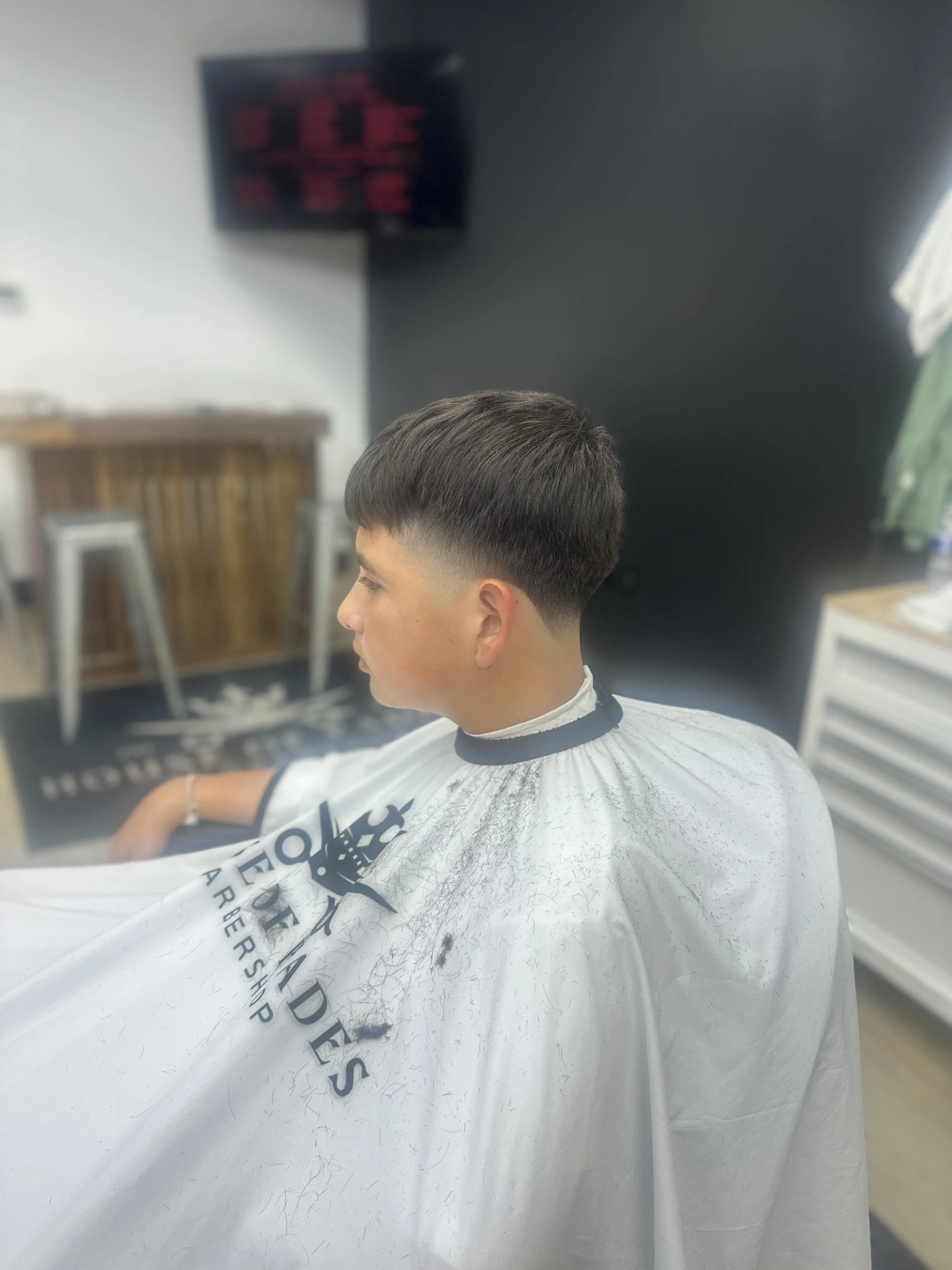 Young man with a short, layered haircut sitting in a barbershop chair, wearing a white barber cape, with a blurred background of barber tools and furniture.
