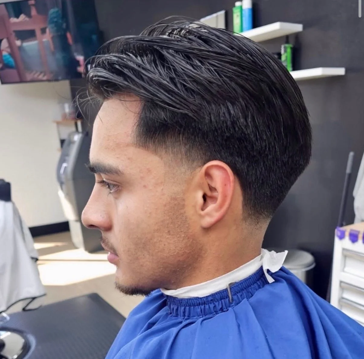 Young man with a freshly styled haircut, featuring a fade on the sides and slicked back hair on top, sitting in a barbershop.