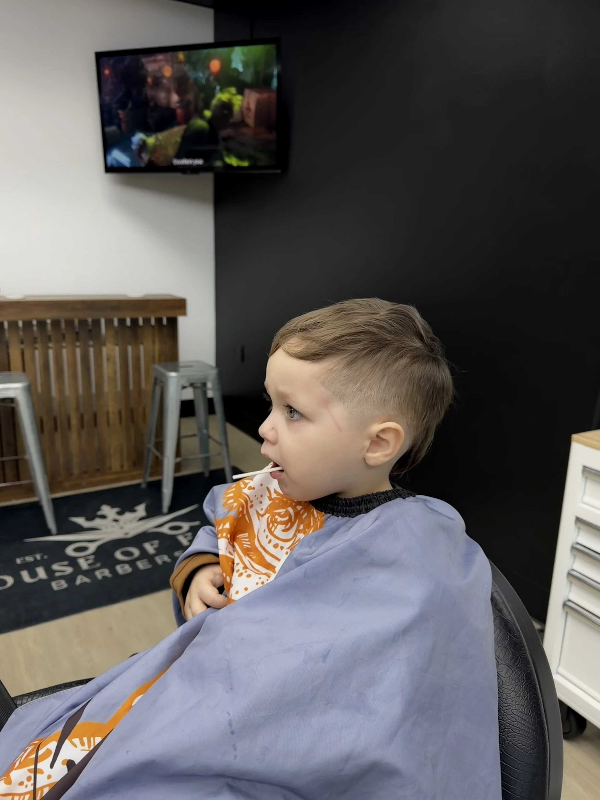Young boy sitting in a barber's chair at a barbershop, wearing a haircut cape, watching TV mounted on a black and white wall.