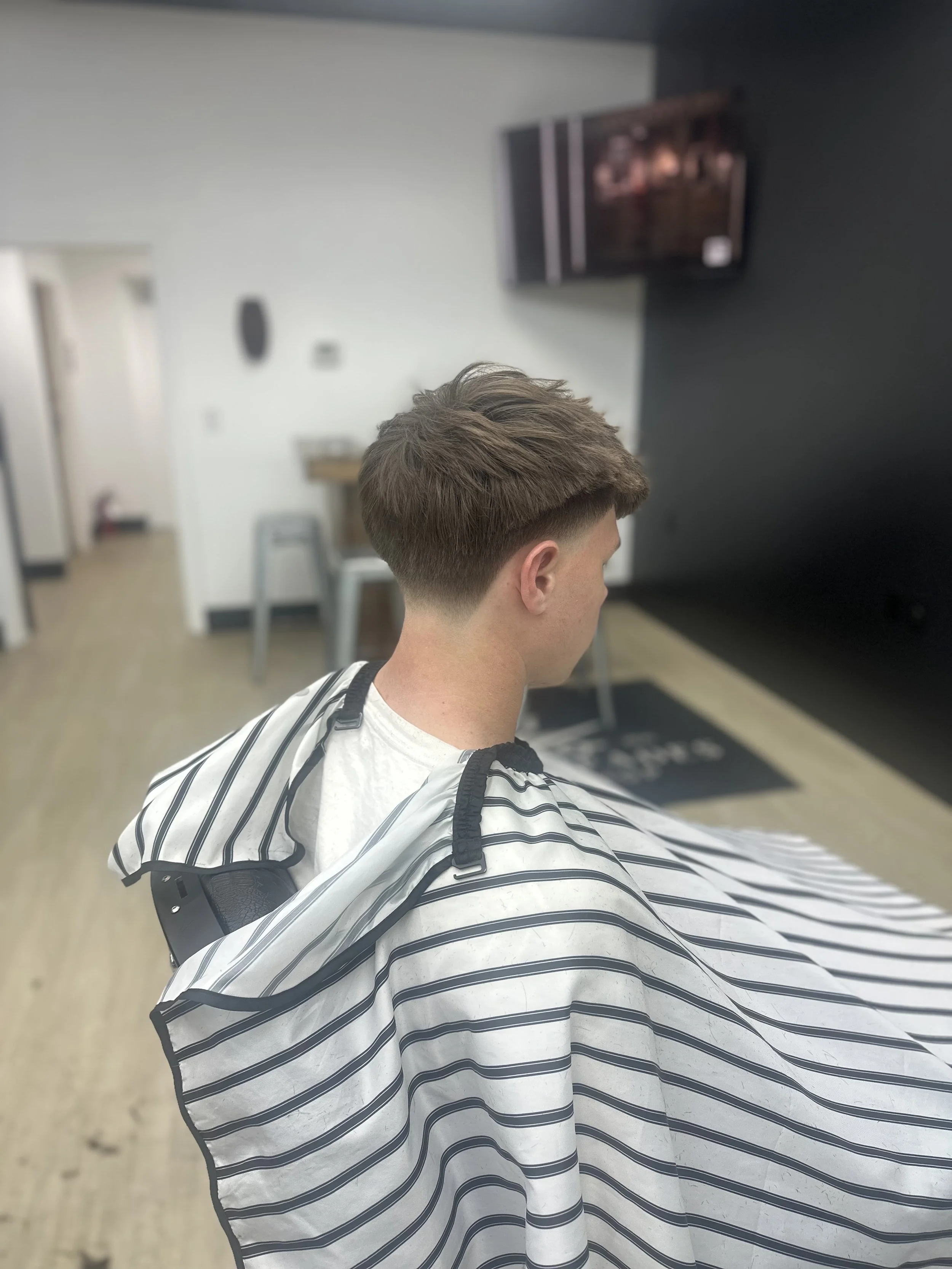 A young man with light skin getting a haircut in a modern barbershop, sitting on a chair with a striped cape draped over his shoulders. The background features a television mounted on the wall and some furniture.