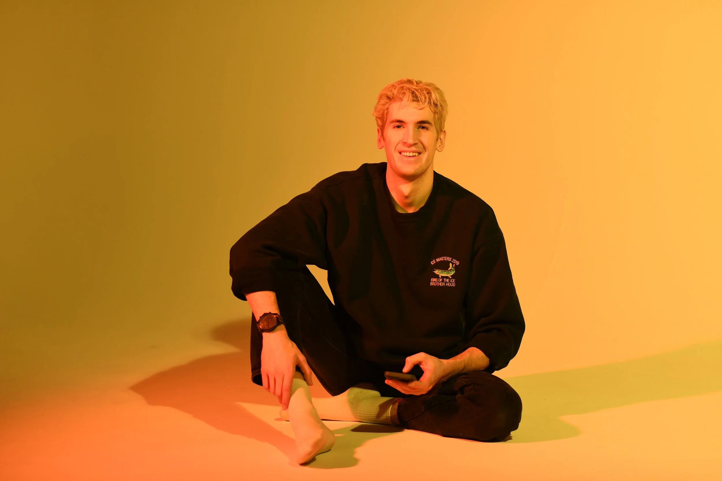 Hunter Cairns is sitting on the floor with a colourful gradient background, smiling, wearing a black sweatshirt,  and holding a smartphone.